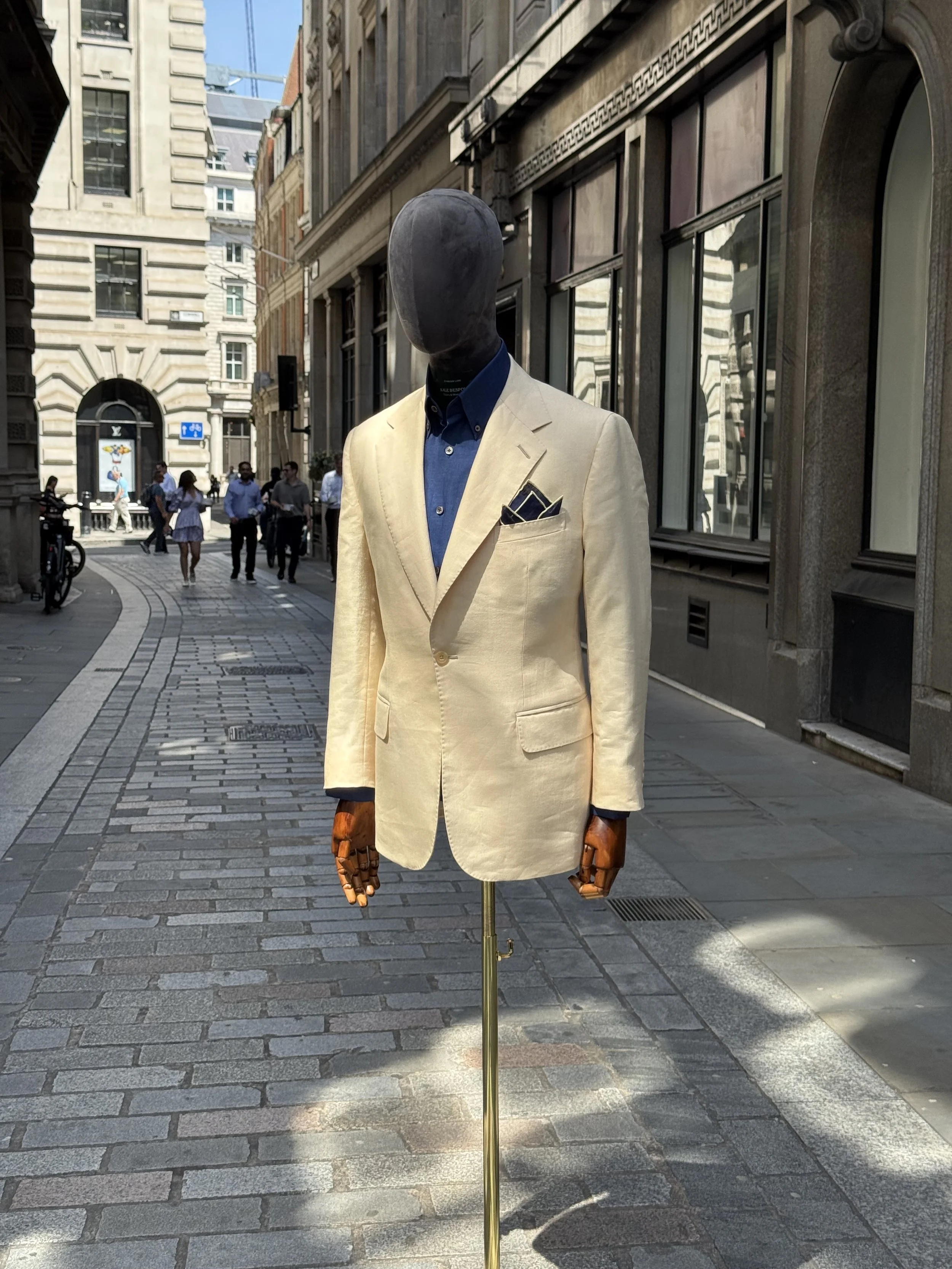 A mannequin dressed in a beige blazer with a blue shirt and pocket square, set up on a city street with buildings and pedestrians in the background.