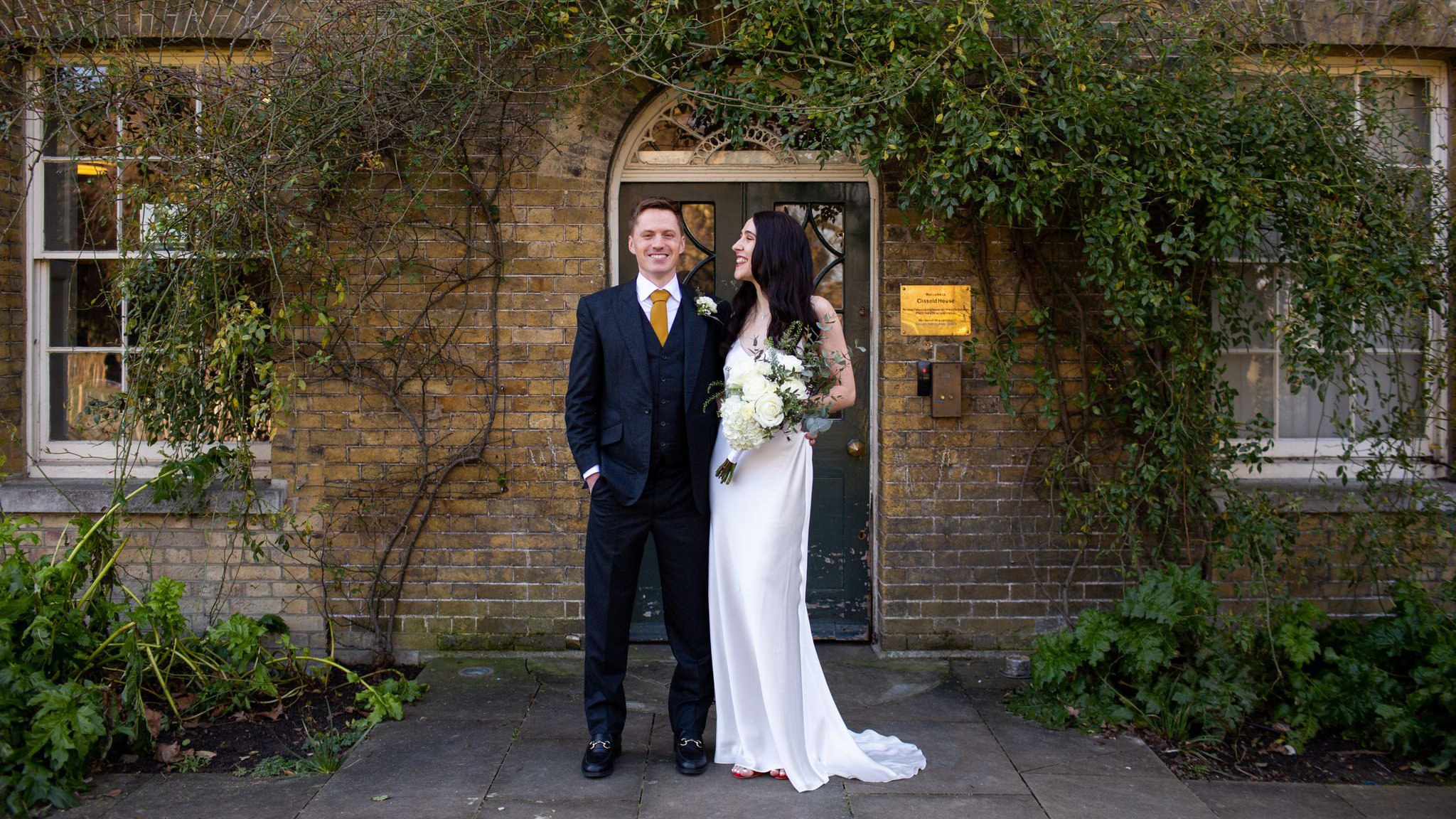 A newlywed couple standing in front of a brick wall with greenery. The groom is wearing a dark suit with a white shirt and yellow tie, smiling. The bride is in a white wedding dress holding a bouquet of white flowers, looking at the groom and smiling.
