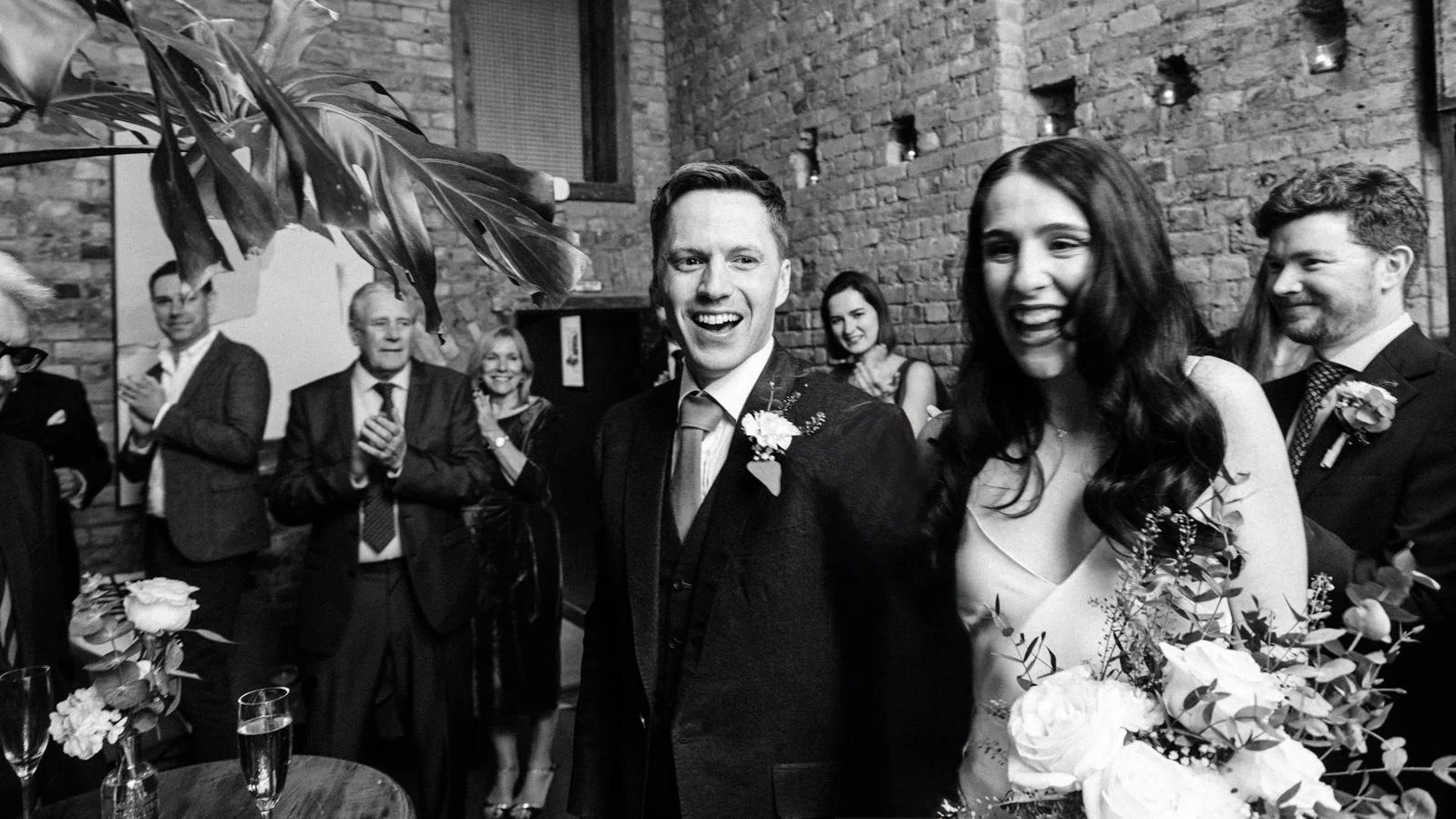 A smiling couple dancing at their wedding reception, surrounded by happy guests in a brick-walled venue.