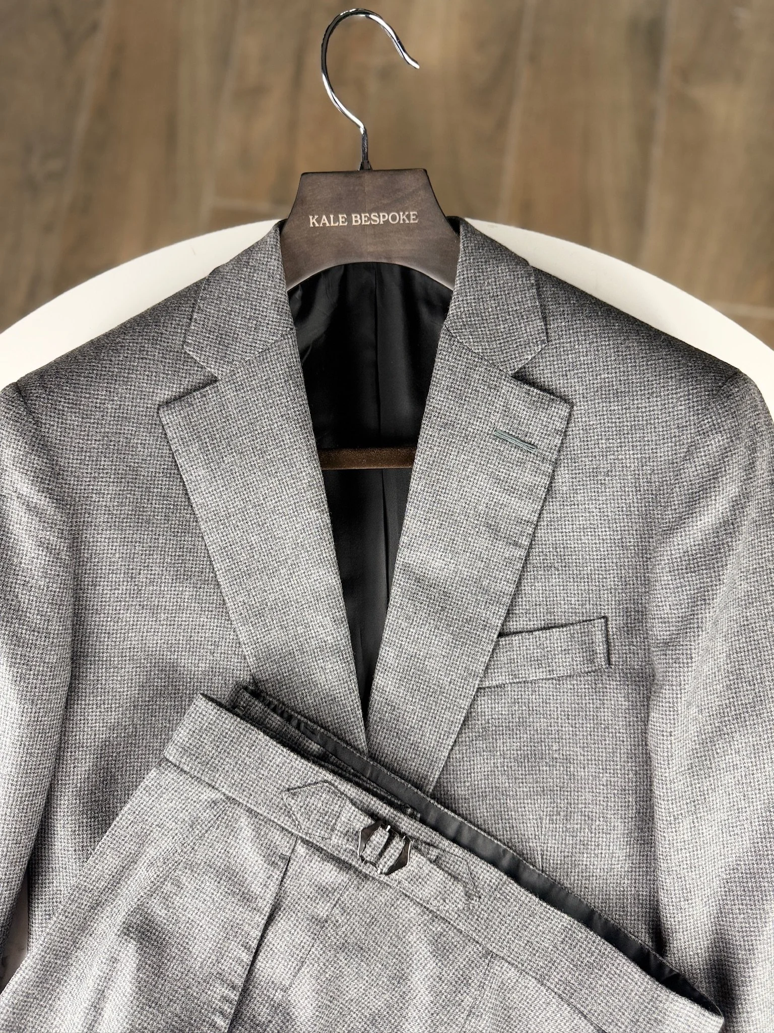 Gray houndstooth men's suit jacket and matching trousers hanging on a wooden hanger with the label 'KALE BESPOKE,' on a white round table, against a wooden background.