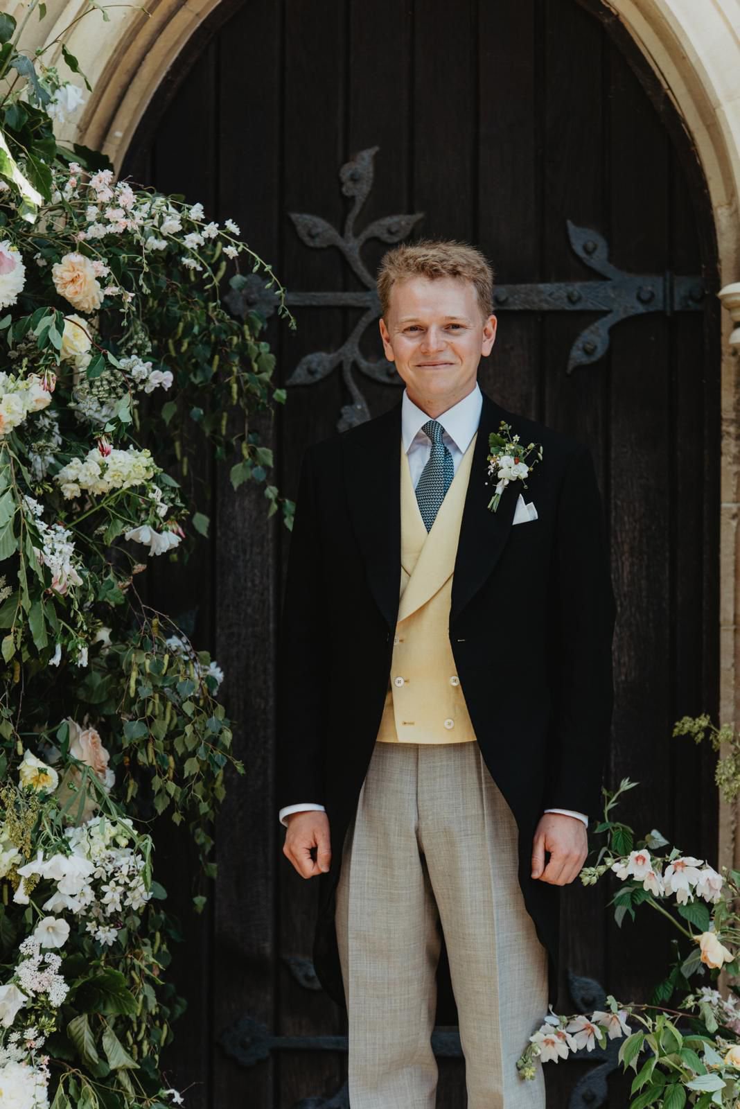 A man dressed in formal attire, including a black coat, cream vest, light-colored plaid trousers, white shirt, and patterned tie, standing in front of a dark wooden door with iron details, surrounded by floral arrangements with white and pink flowers and greenery.