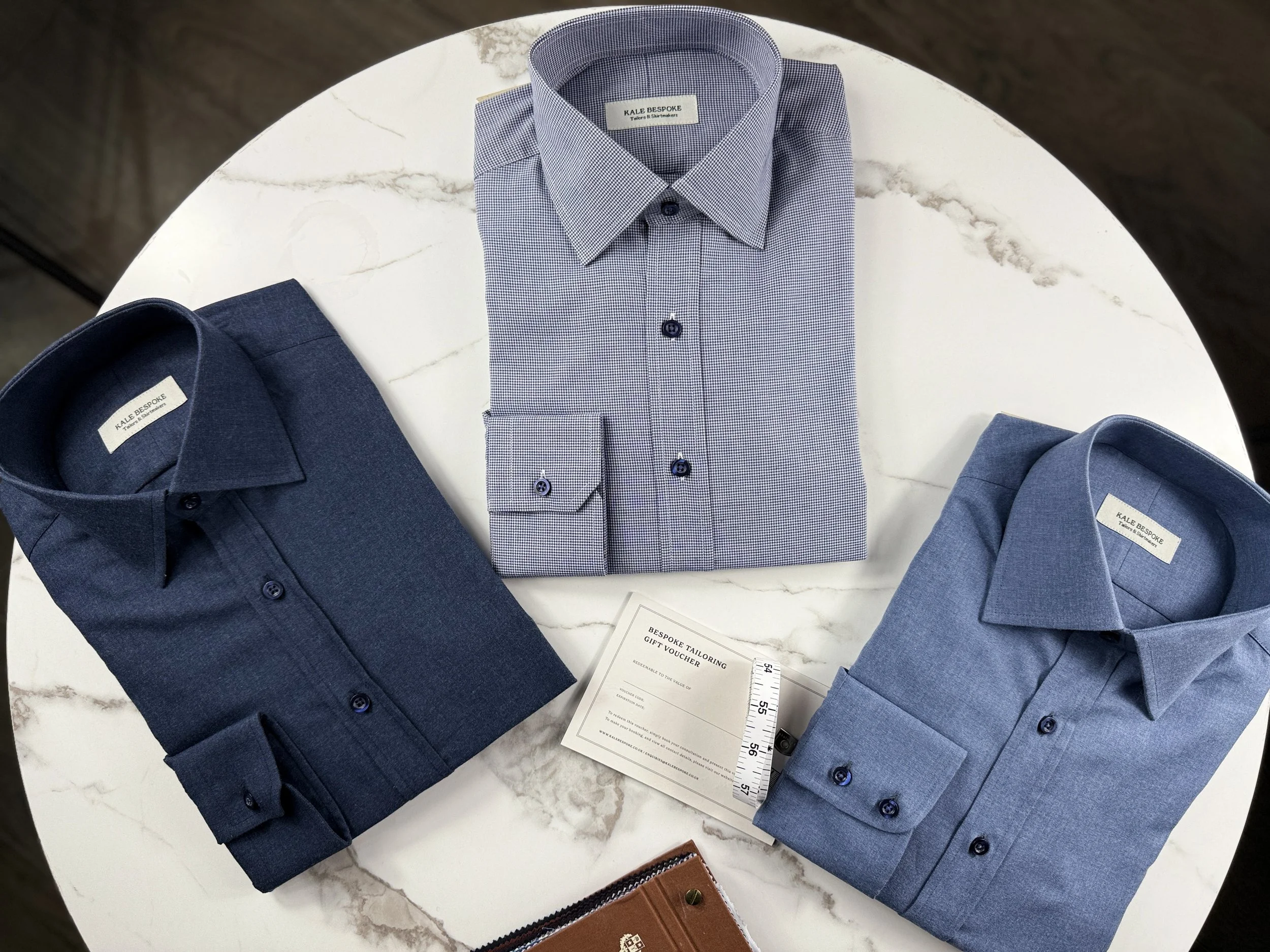 Three folded dress shirts in shades of blue are displayed on a white marble tabletop with a small gift card and a brown leather wallet.