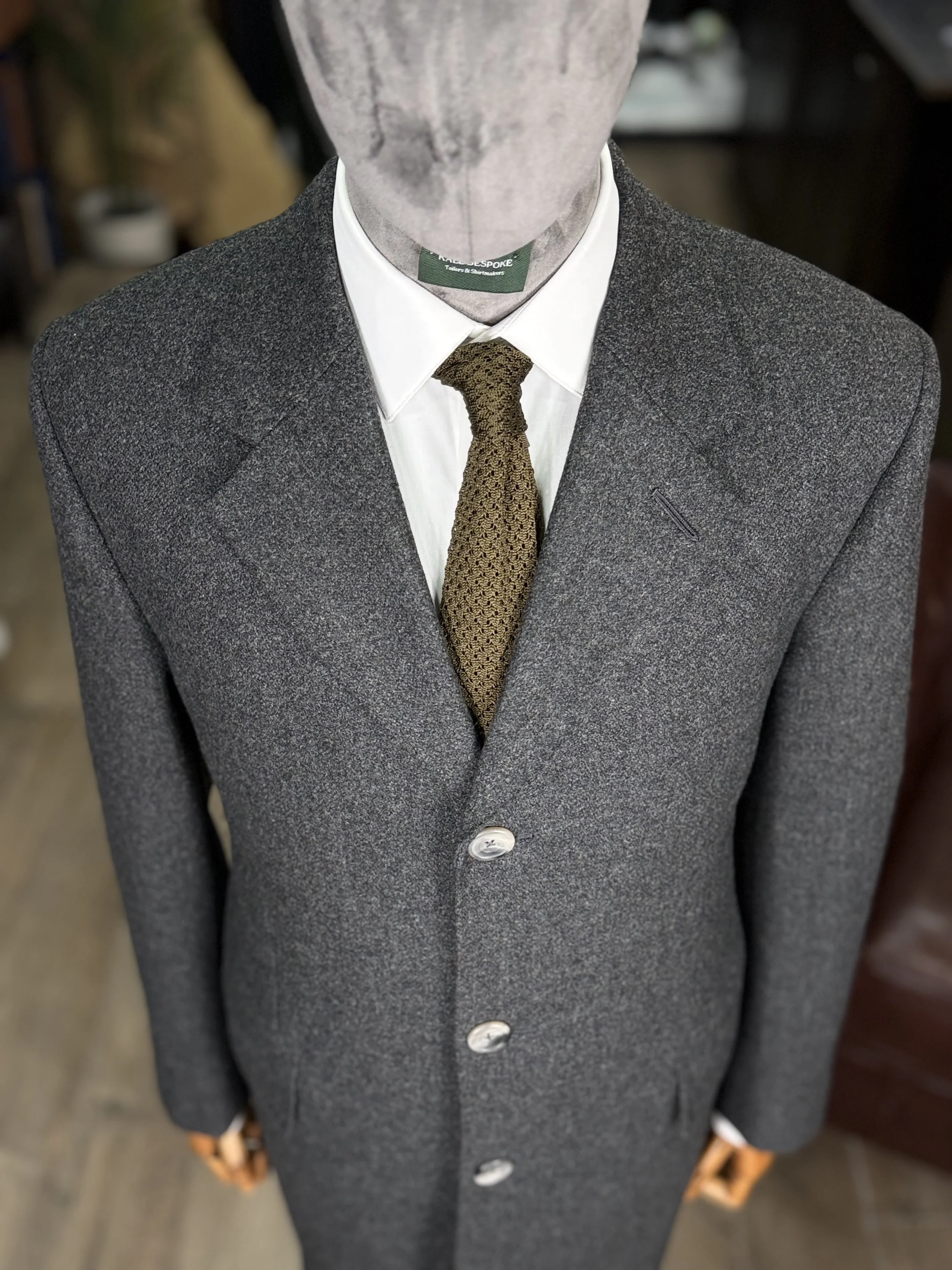 Bespoke Charcoal grey Overcaot No Chest pocket on shop mannequin with a silk knitted green silk tie.This overcaot fabric is 20oz Drim dugdale brothers full hand cut and handmade Kale bespoke Overcoat.Made by Kale Bespoke