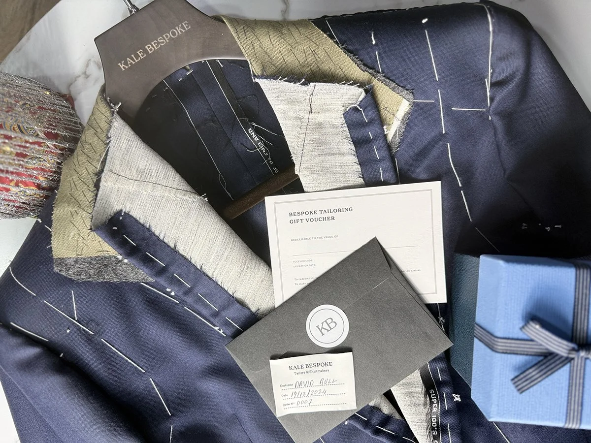A collection of fabric swatches from KALE BESPOKE, a gift voucher for bespoke tailoring, a black KALE BESPOKE shoebox, and a blue gift box with a black ribbon.