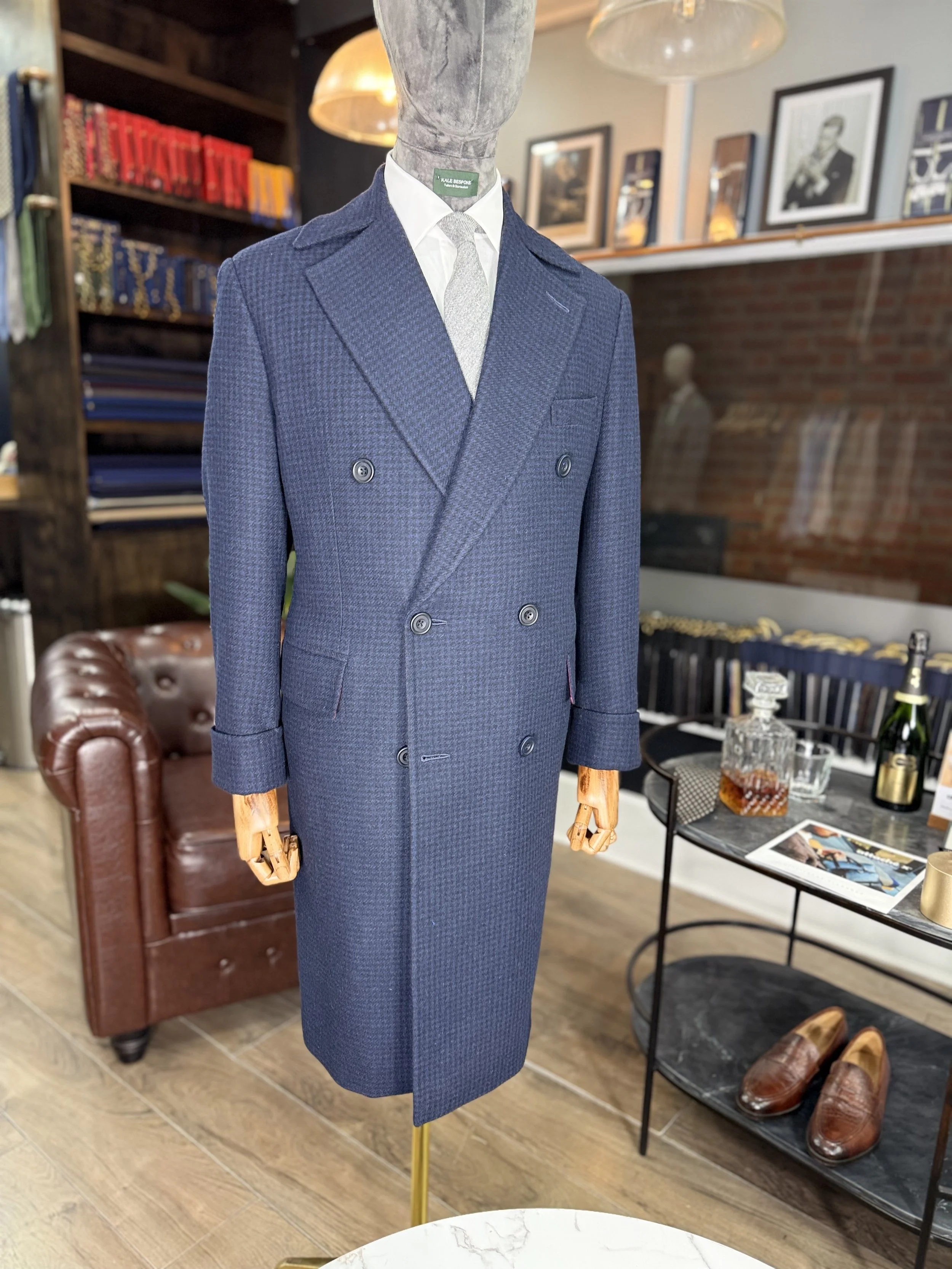 A mannequin dressed in a double-breasted navy blue suit with a white shirt and light-colored tie. The setting appears to be a vintage or upscale clothing store with books, framed art, a leather armchair, a marble-top table with drinks and glasses, an