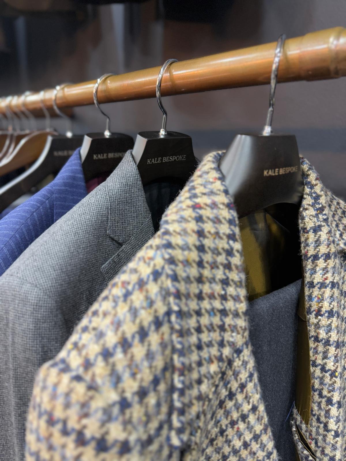 A clothing rack with jackets and blazers on hangers labeled 'KALE BESPOKE,' including a blue blazer, a gray blazer, and a patterned blazer with yellow, blue, and white colors.