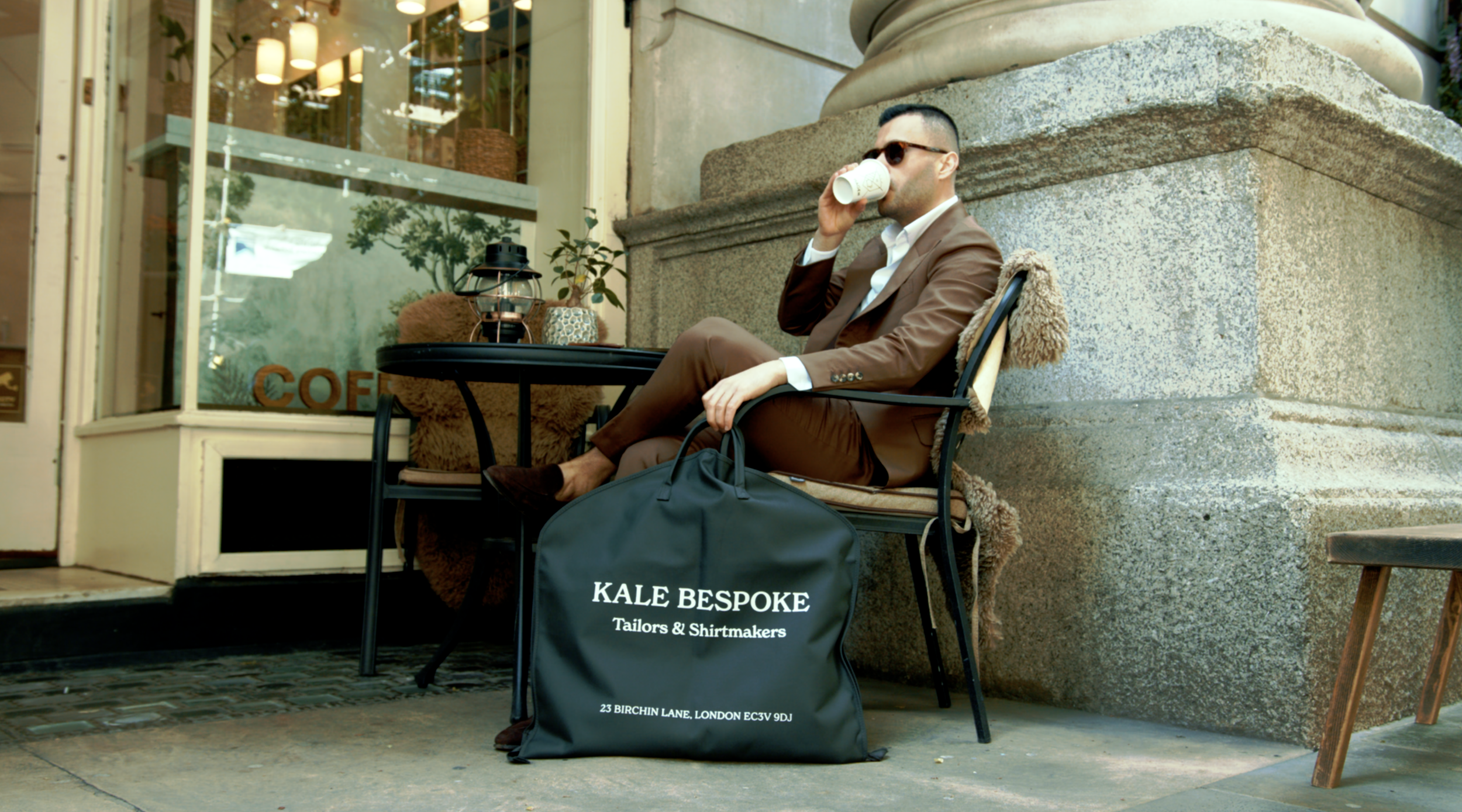 A man in a brown suit and sunglasses sitting outside a café, drinking from a cup, with a large black bag labeled 'KALE BESPOKE Tailors & Shirtings' on the ground next to him.