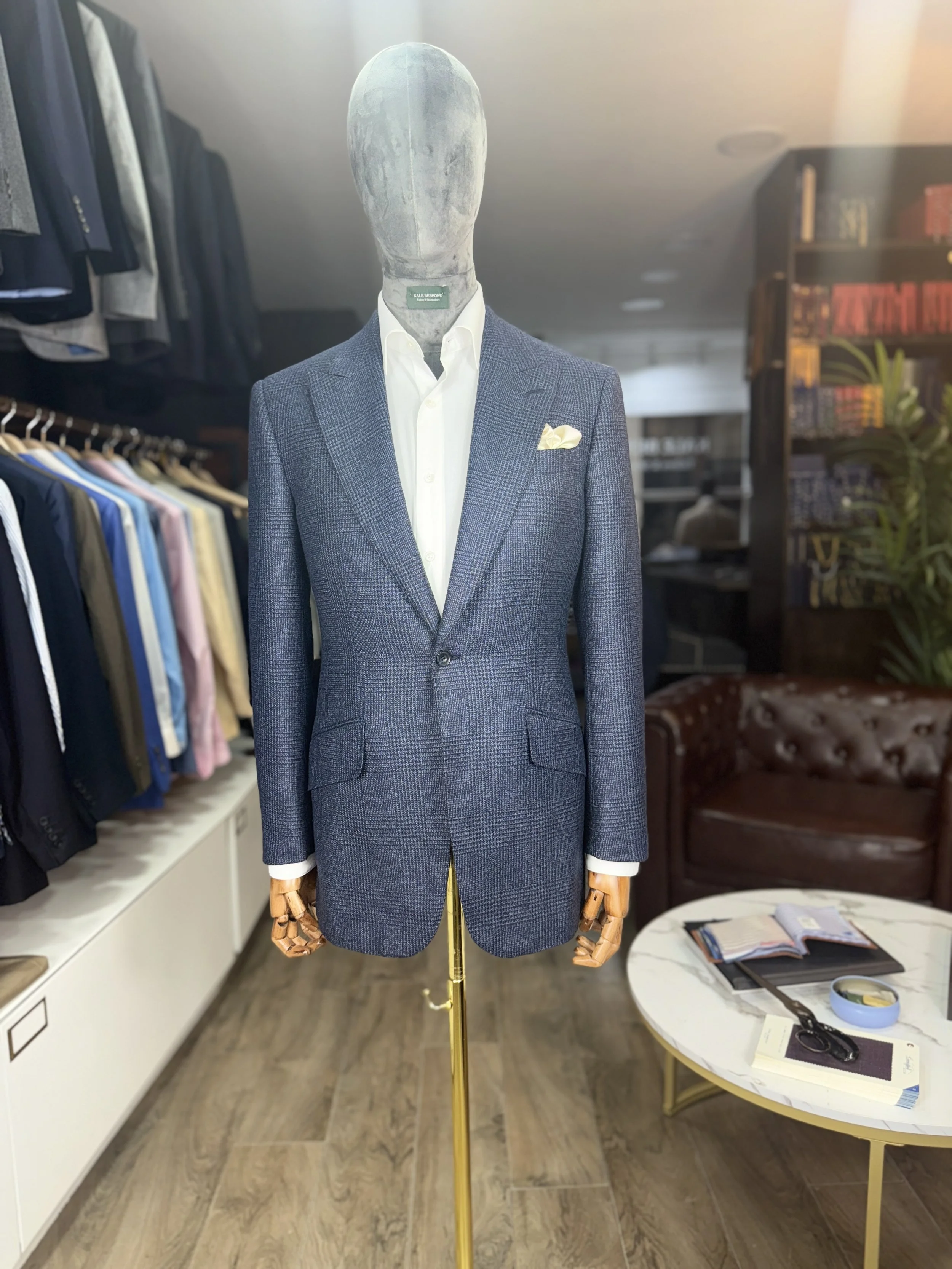 A mannequin dressed in a white shirt and a blue checkered blazer, standing in a clothing store with racks of clothes and a bookshelf in the background.