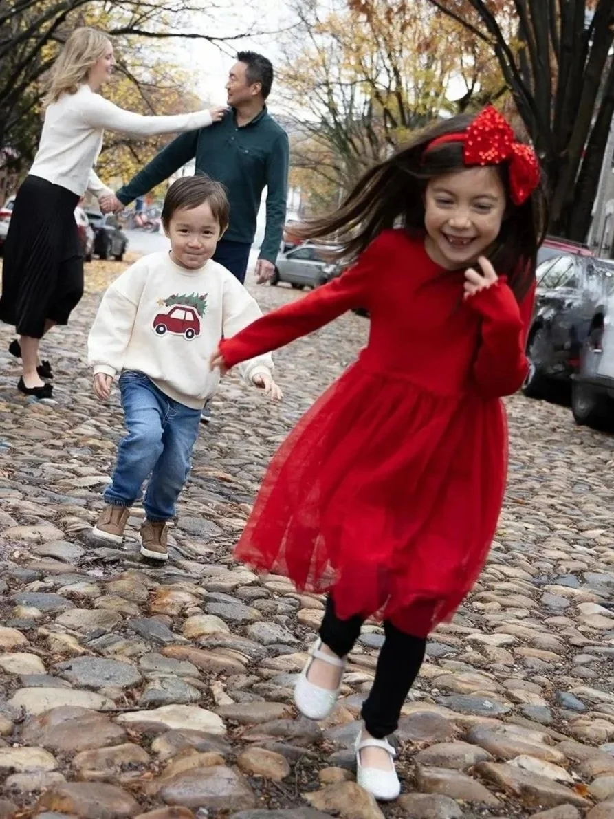 Children running on cobblestone street, girl in red dress with bow smiling, boy in white sweater with car and Christmas tree design, adults holding hands in background, trees with fall foliage.
