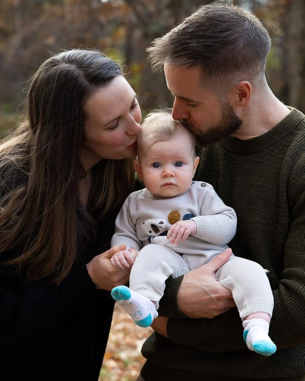 A family of three outdoors in autumn. The mother and father are kissing their baby on the forehead. The baby looks directly at the camera with a serious expression, wearing a cream-colored outfit with colorful polka dots and white socks with teal accents.