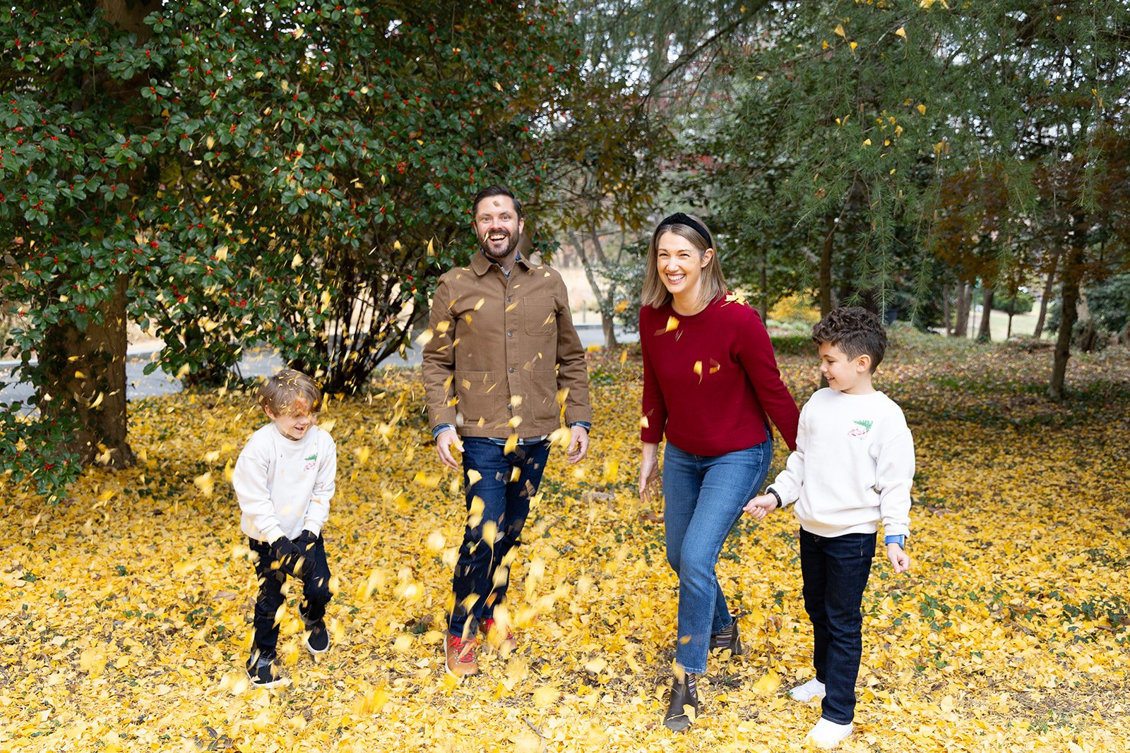 A family of four, two parents and two children, happily playing in a park filled with yellow leaves during autumn, with trees in the background.