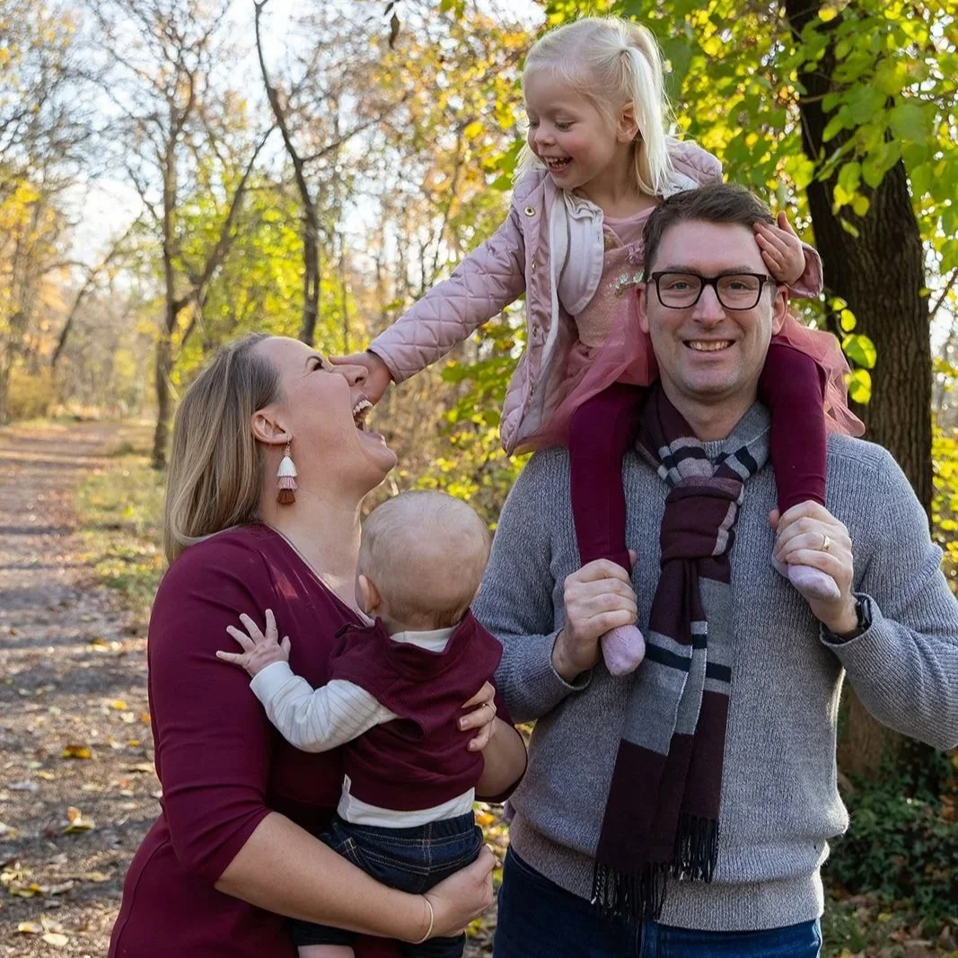A family of four enjoying a walk outdoors in a park during fall. The mother is holding a baby, the father is carrying a young girl on his shoulders, and they are all smiling and laughing.
