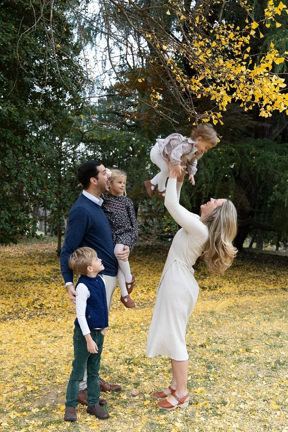 A family of five enjoying fall outdoors, with yellow leaves on the ground and trees in the background. The mother lifts a young girl into the air, while the father holds a young boy and watches. The girl smiles as she is raised, and the father and mother are happy.