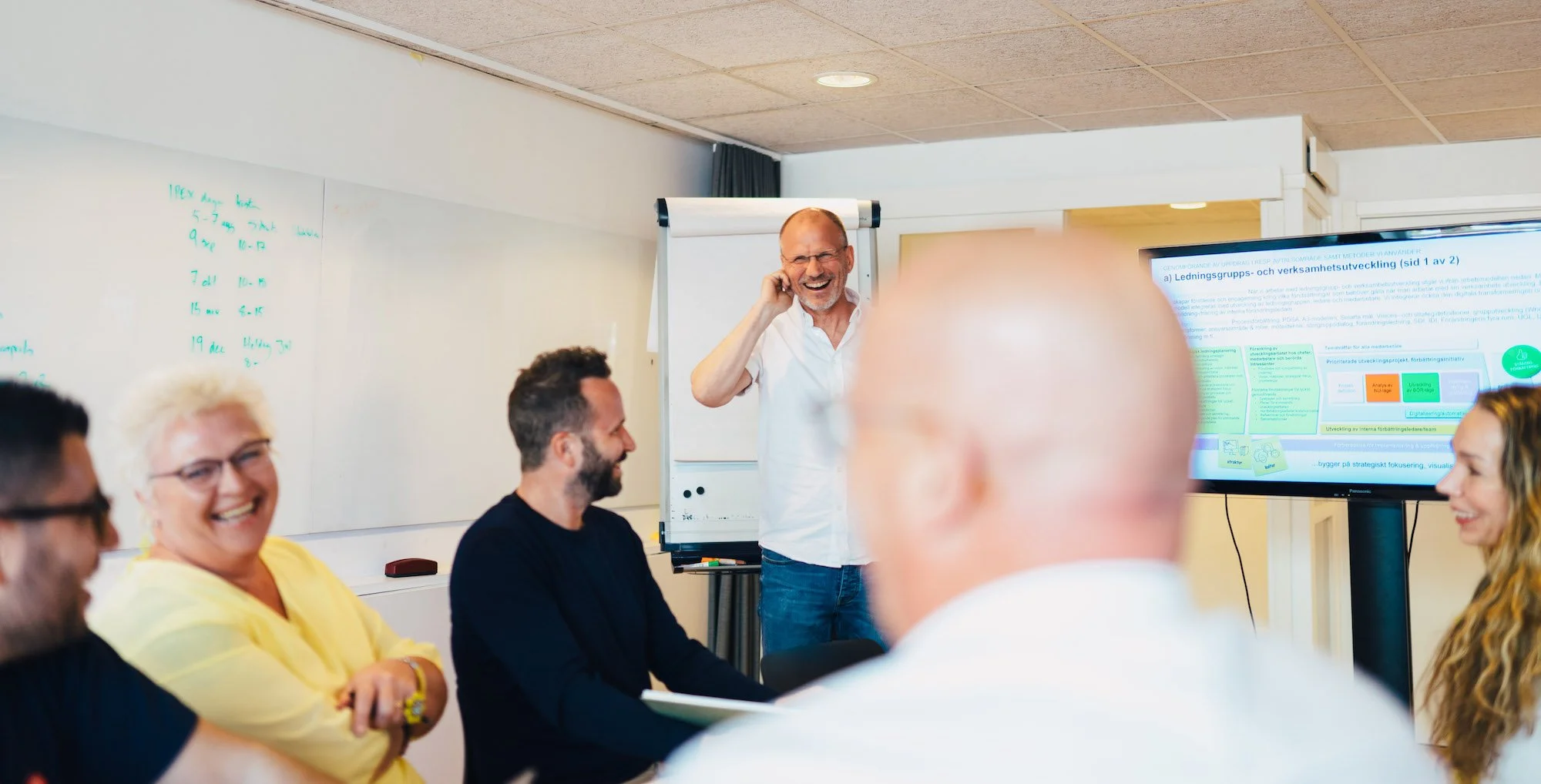 Mänsklig grupp i ett konferensrum, en man står framför en whiteboard och ett stort digitalt skärm, alla andra sitter och ler.