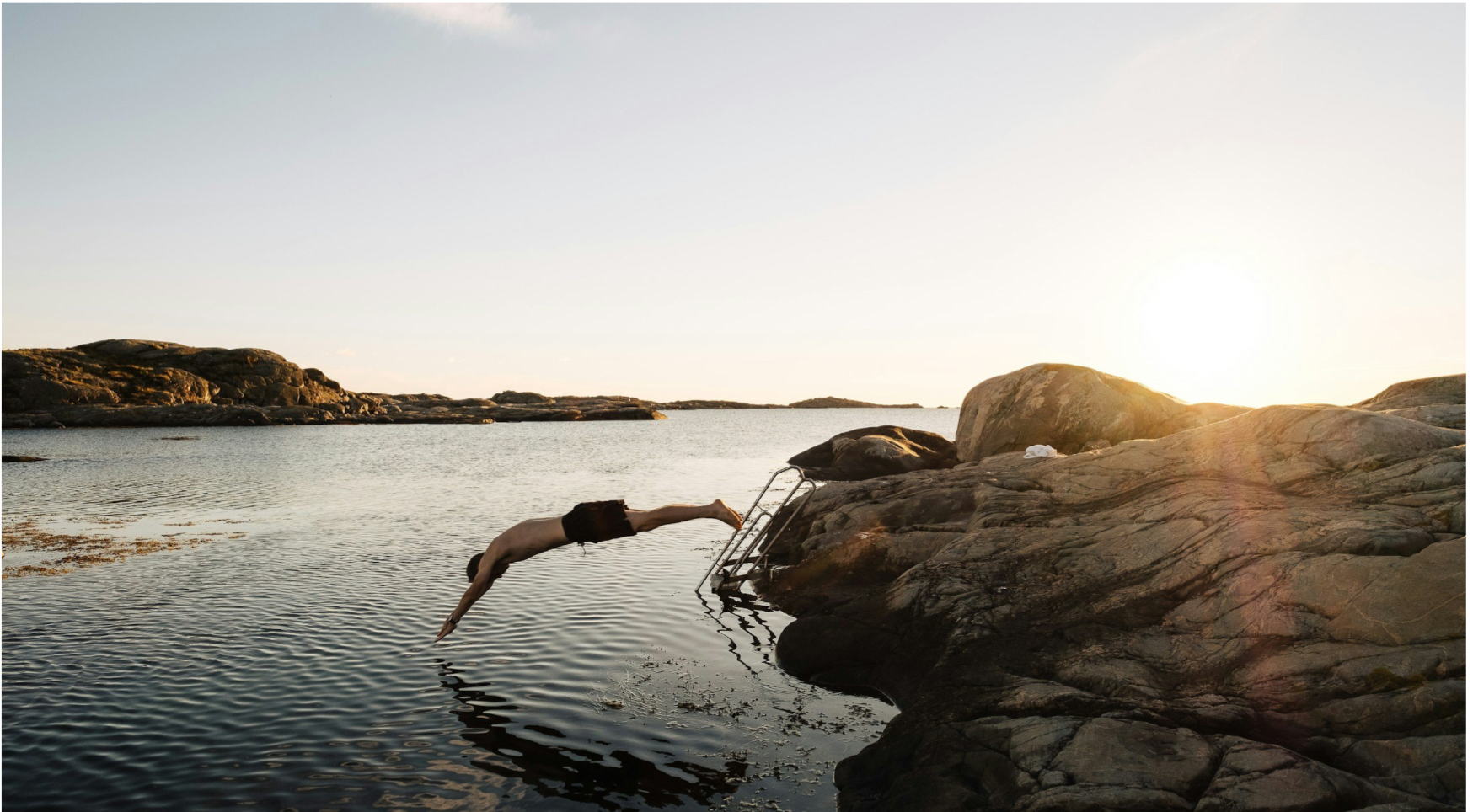 En person hoppar från en klippa ner i vattnet vid solnedgången.