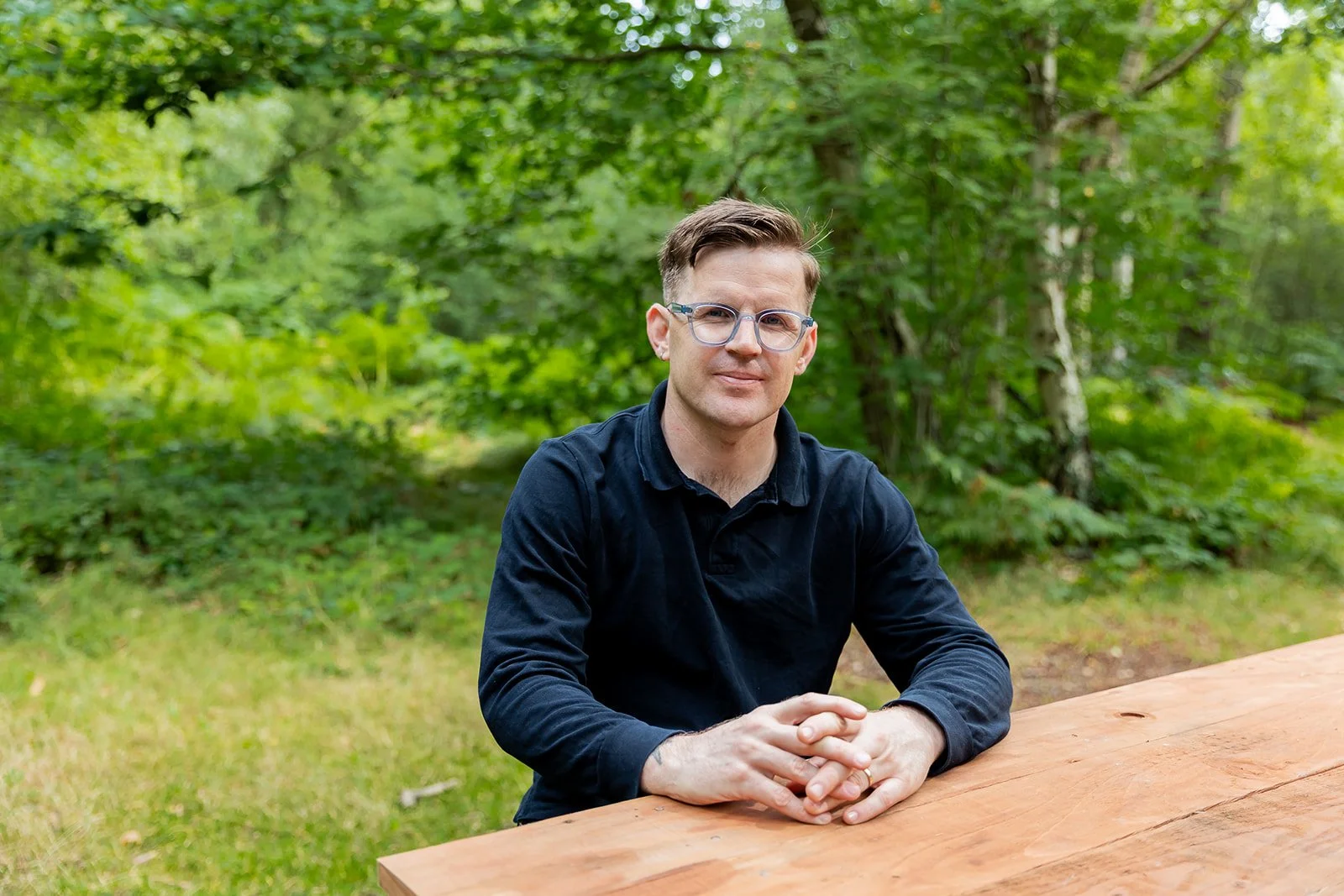 A man with glasses wearing a black shirt sitting at a wooden table outdoors with green trees and grass in the background.