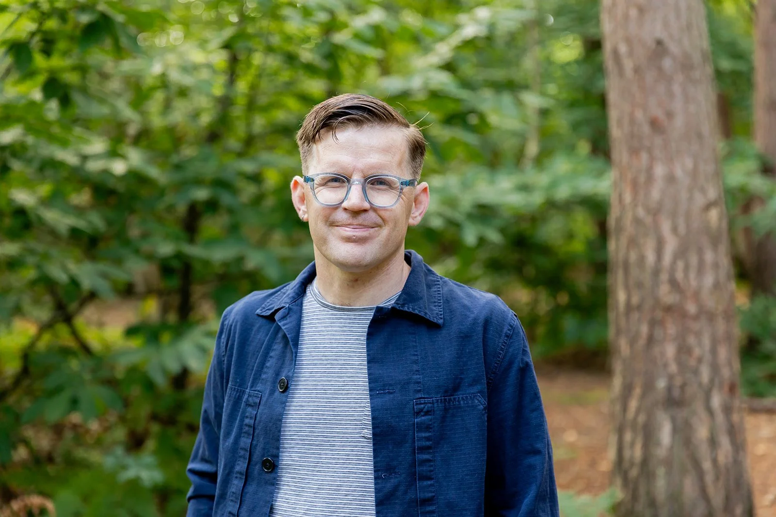 A man wearing glasses and a navy jacket standing outdoors in a forest with green foliage and trees.