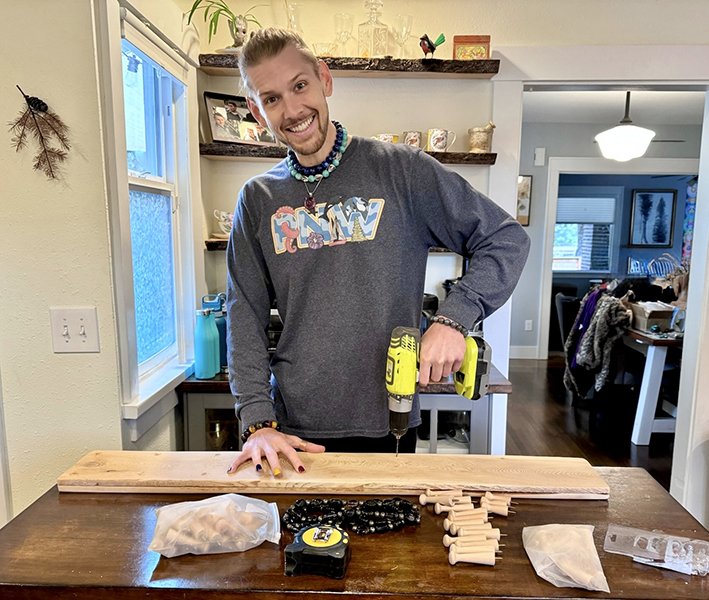 A man smiling at the camera while holding a yellow cordless drill in a kitchen, next to a woodworking project with small wooden dowels, black beads, and a plastic bag on the table.