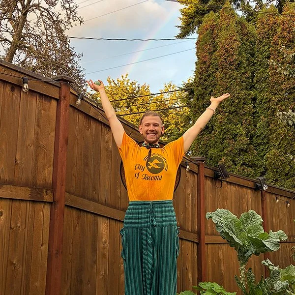 A man standing in a backyard with a wooden fence, smiling with arms raised, wearing an orange T-shirt with a fish logo and the words 'Aqu Affair of Tacoma,' blue striped pants, and a black mask around his neck. There is a rainbow visible in the sky, and trees with green foliage in the background.