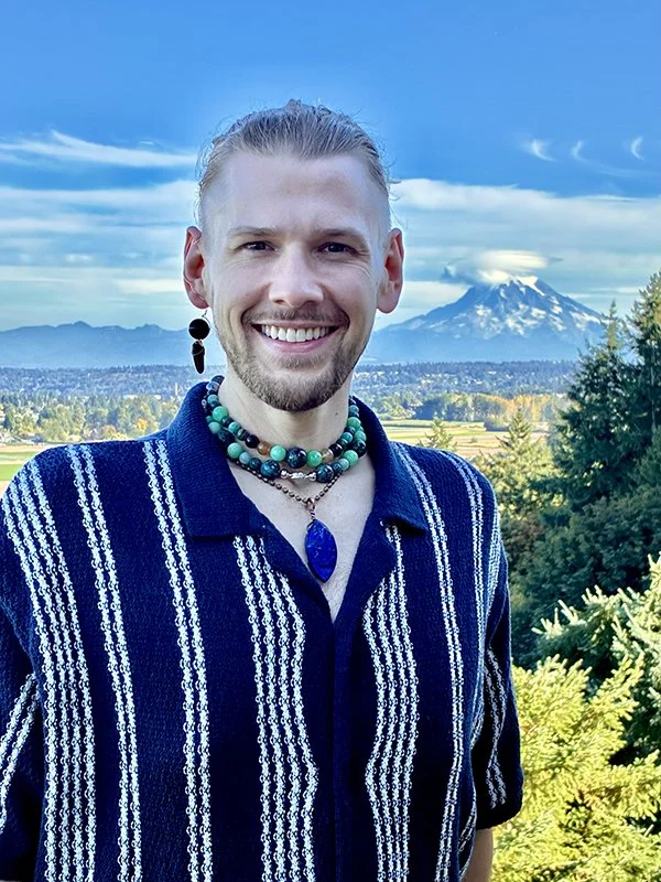 A smiling man with long hair tied back, wearing a striped blue and white shirt with beaded necklaces and earrings, standing outdoors with a mountain and blue sky in the background.