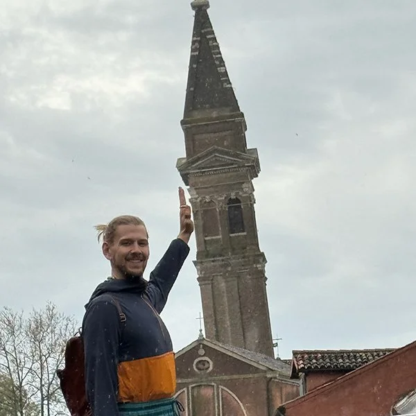 A man smiling and raising his hand in front of a tall church steeple.