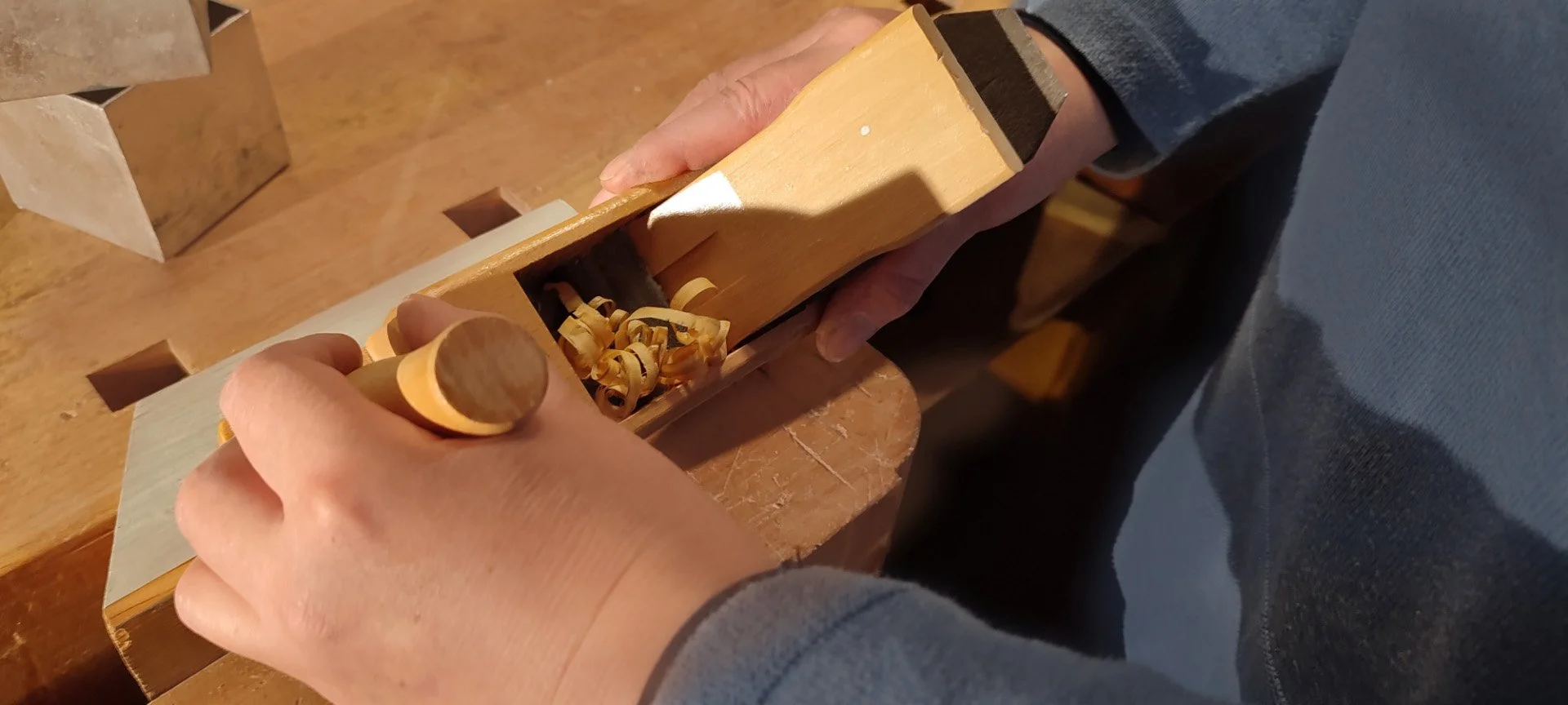Person hobelt Holz mit einer Handhobelmaschine, Holzspäne fallen ab, auf einem Tisch