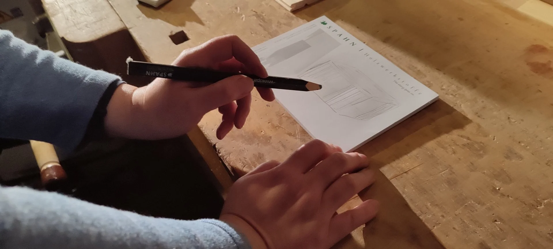 Person schreibt auf einem Notizblock mit einem Bleistift, der auf einem Holztisch liegt.