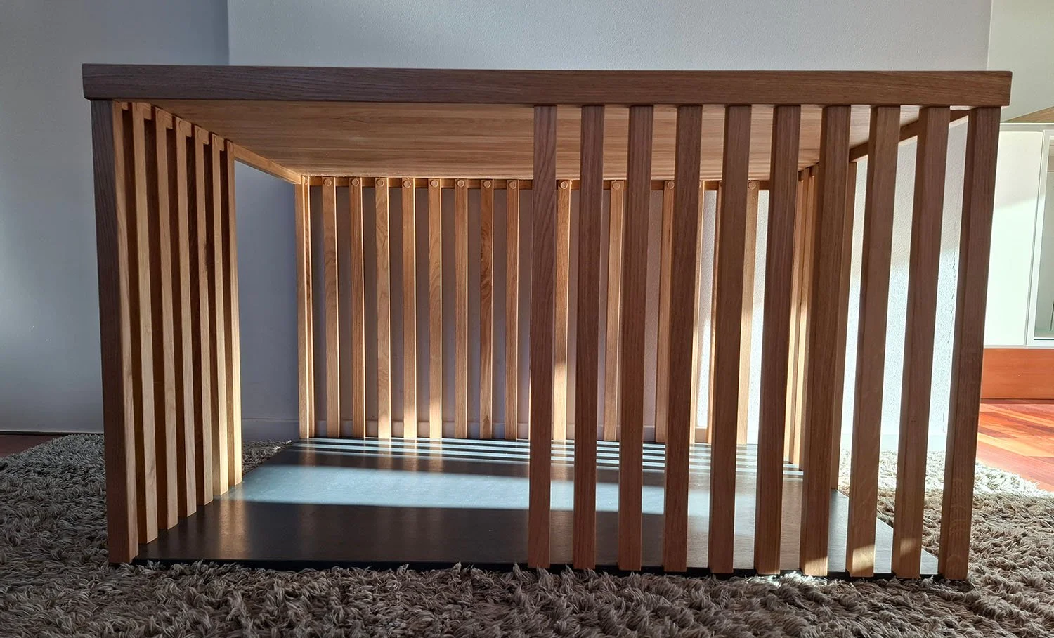 Eine Hundebox aus Holz mit vertikalen Lamellen steht auf einem Teppichboden in einem Raum.