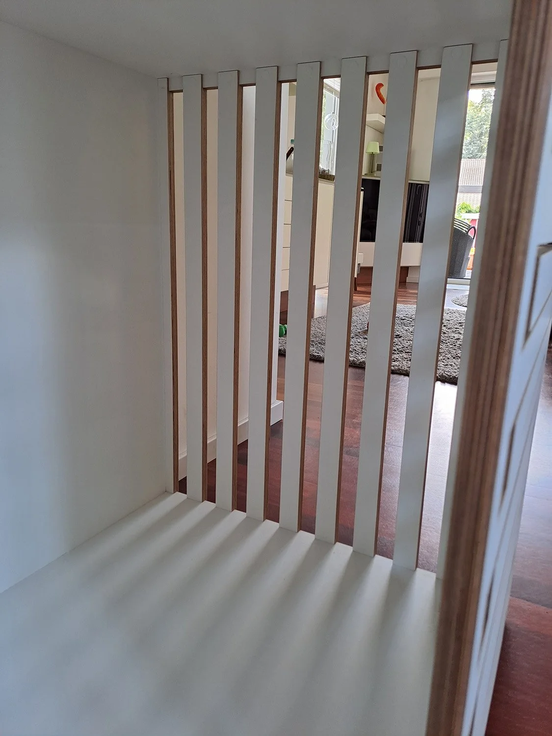 Ein Blick durch eine kleine weiße Hundebox aus Holz in einem Wohnzimmer mit braunem Holzboden, einem grauen Teppich und Balkon mit Pflanzen im Hintergrund.