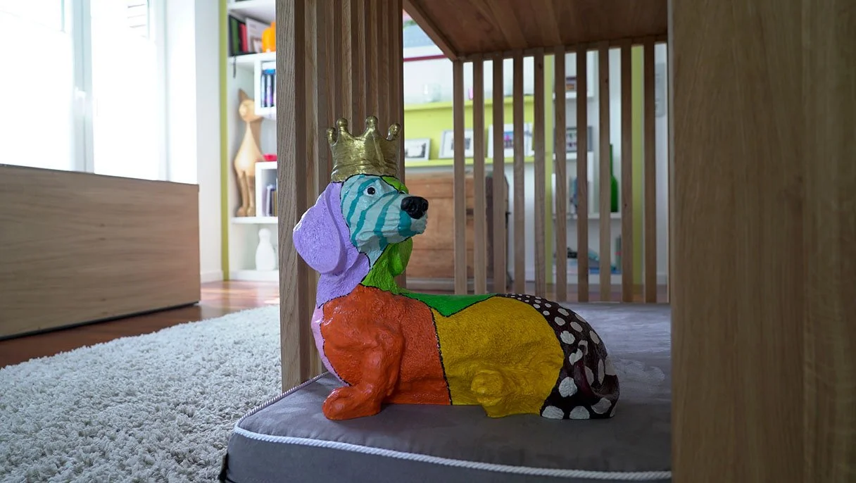Eine bunte Hundefigur mit Krone, die auf einem Kissen unter einem Holztor sitzt.