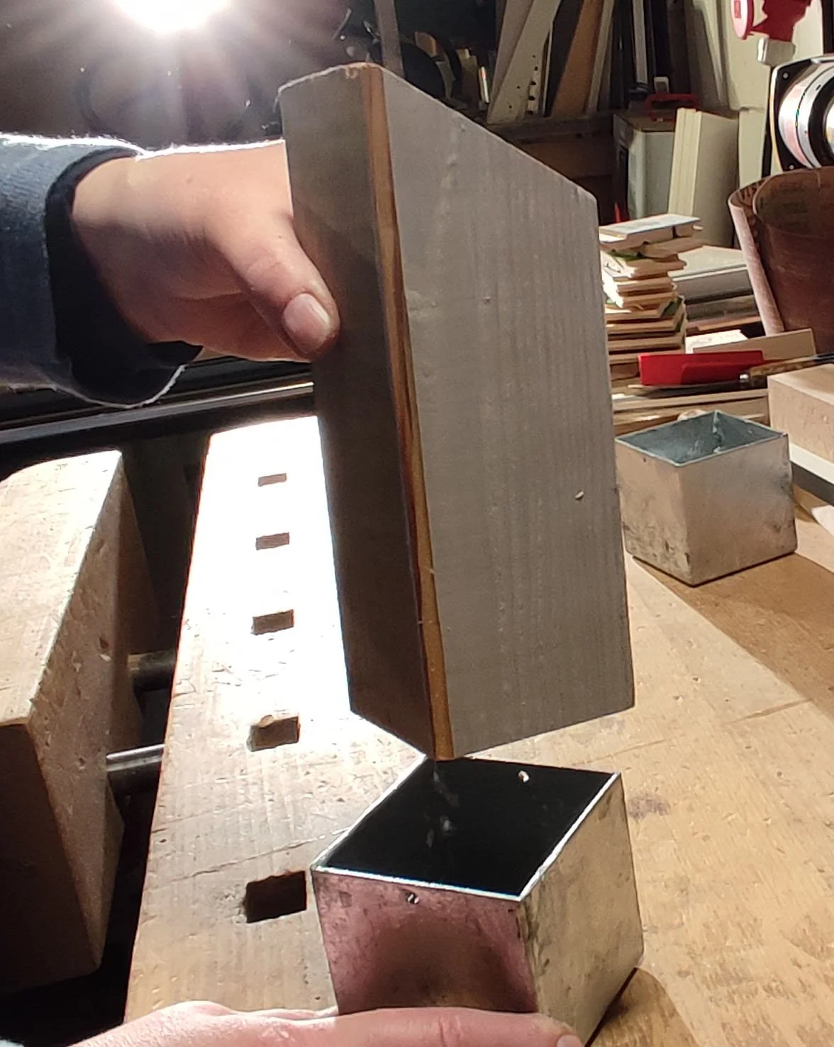 Jemand hält ein Stück Holz über eine Metallbox in einer Werkstatt. Im Hintergrund sind Werkzeuge und vereinzelte Holzstücke zu sehen.