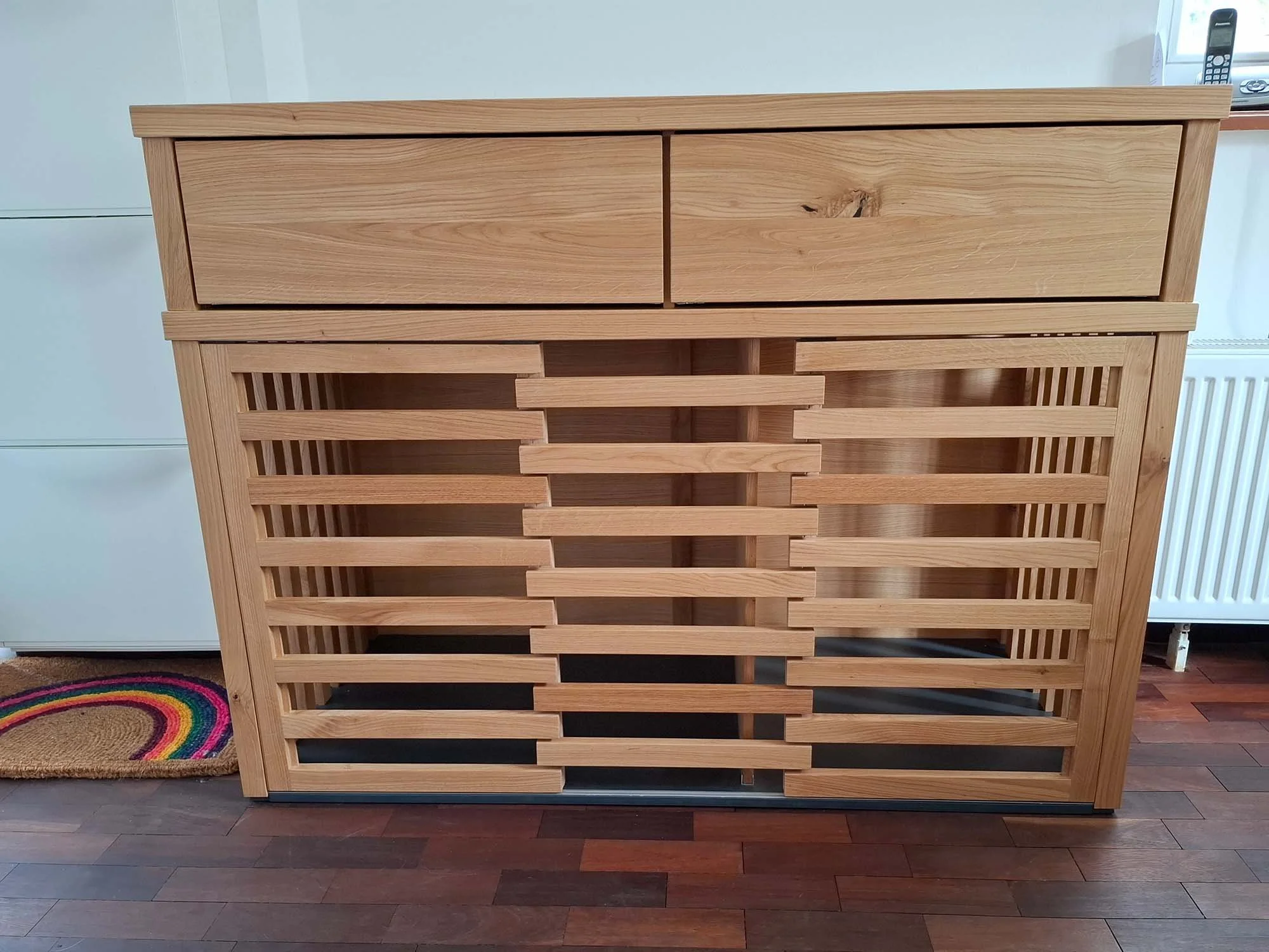Ein moderne Hundebox aus Holz mit zwei Schubladen oben und einer Schiebetür mit horizontalen Lamellen unten, auf einem Holzboden neben einem weißen Möbelstück.