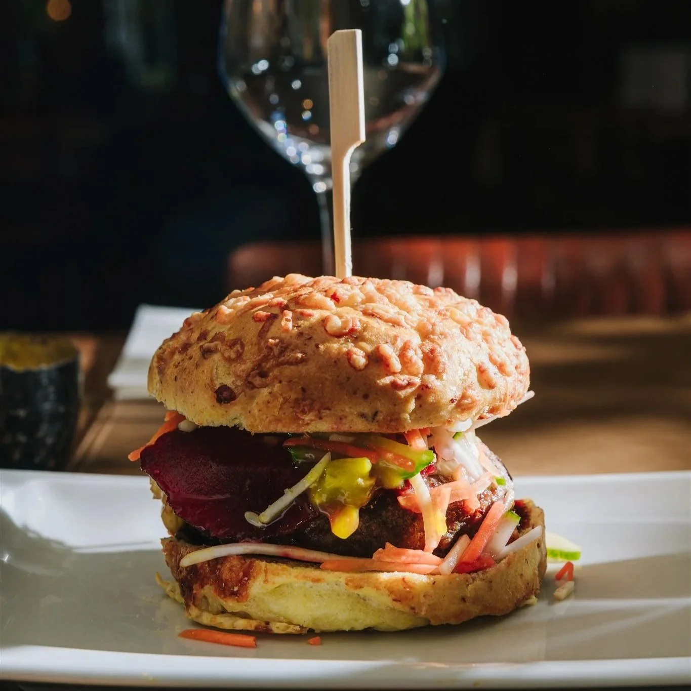 Een goed gevulde hamburger op een wit bord, in de achtegrond staat een wijnglas.