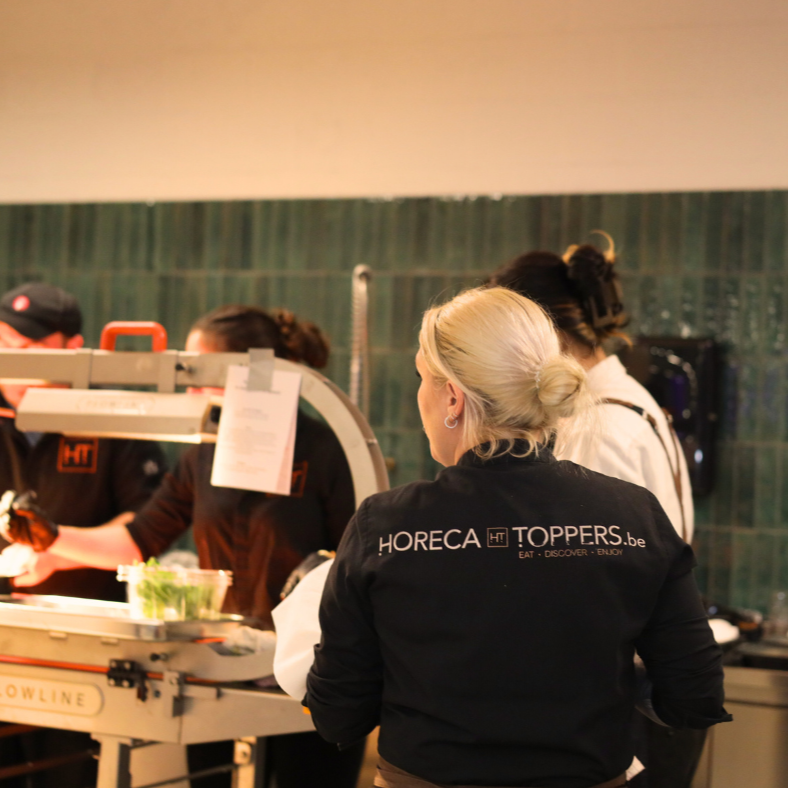 Personeel in een keuken, met één vrouw op de voorgrond met een zwarte jas met de tekst 'HORECA TOPPERS.be'.