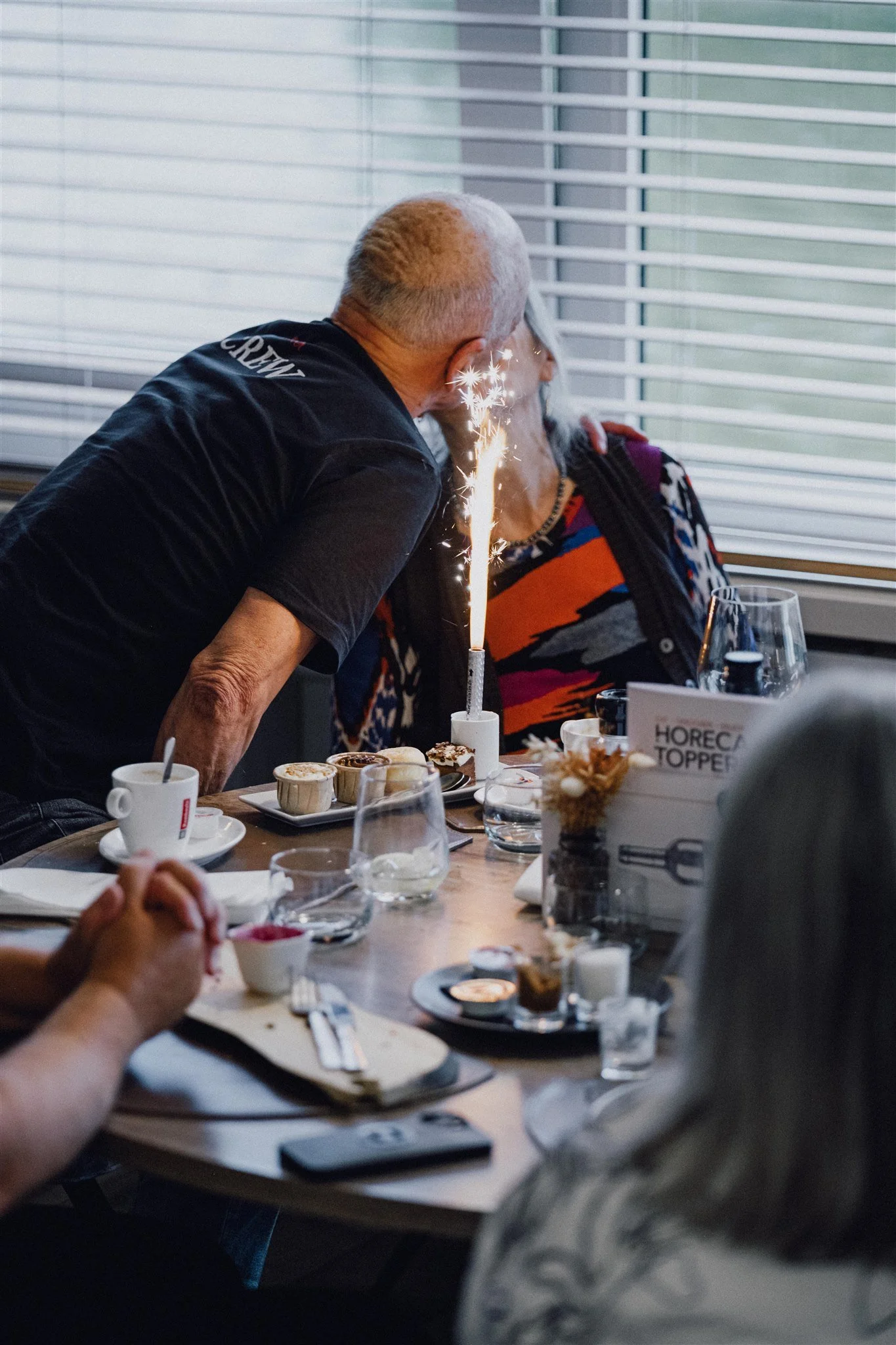 Een man kust zijn vrouw voor haar verjaardag, er brand een vuurwerkje op de voorgrond. De sfeer is feestlijk en liefdevol.