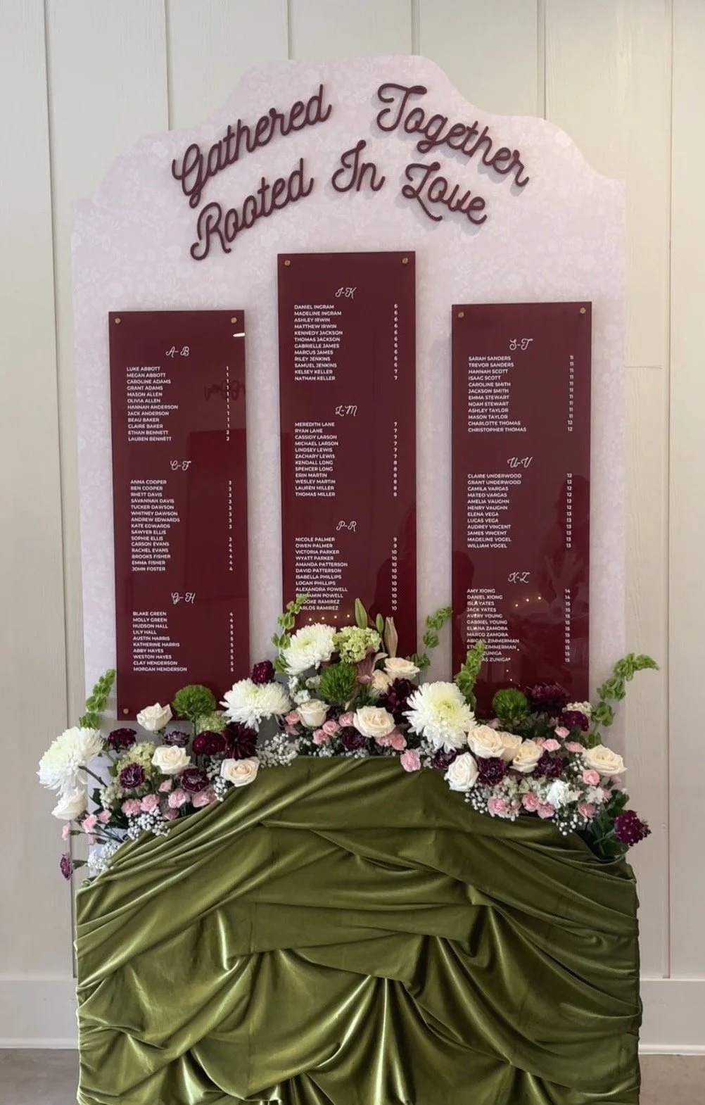 Custom burgundy acrylic wedding seating chart with three personalized panels, lush floral arrangements, and olive green draping, designed and built by Covenant Design Co. for an event at Bella Cavalli in Aubrey, Texas.