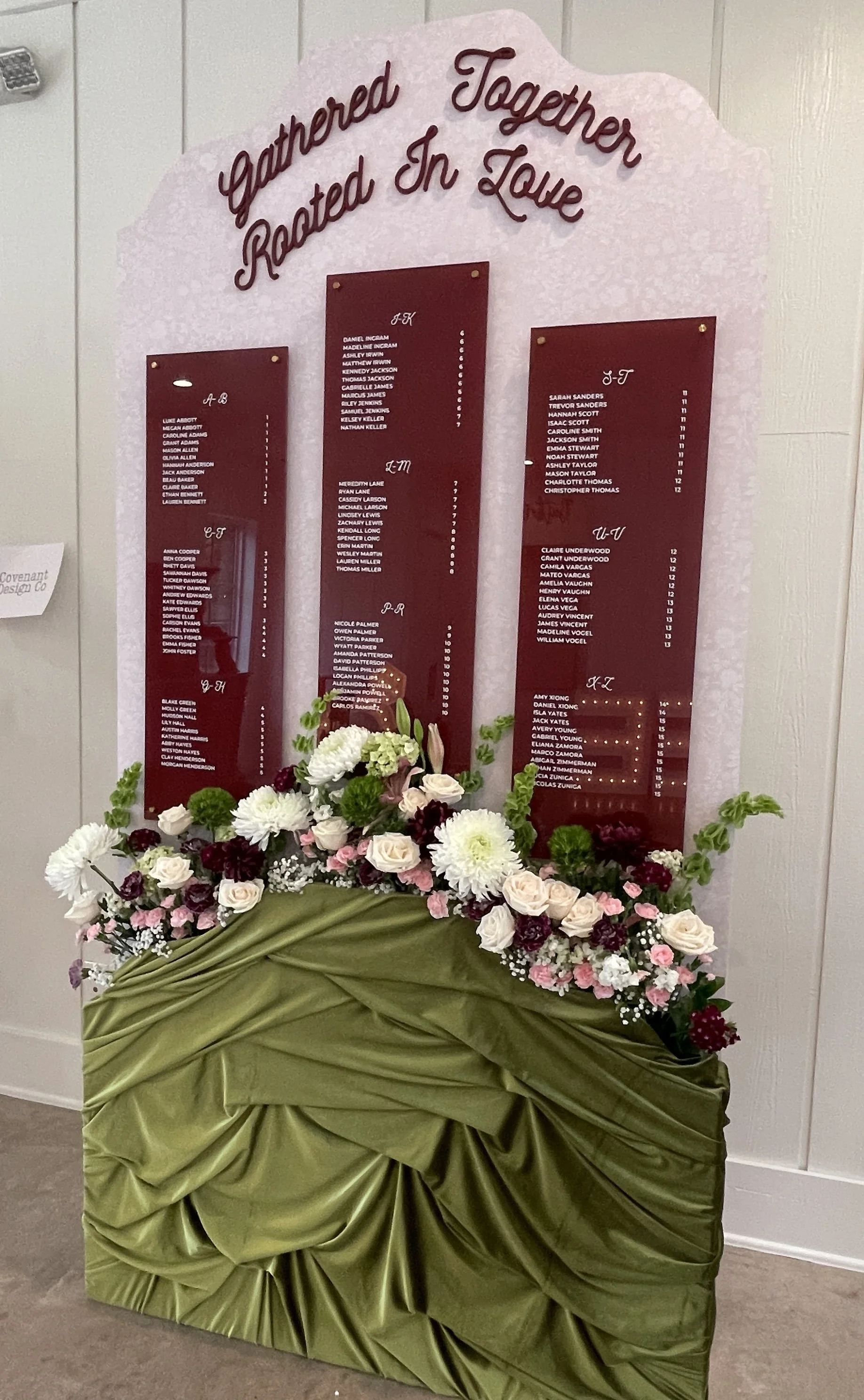 Custom burgundy acrylic wedding seating chart with three personalized panels, lush floral arrangements, and olive green draping, designed and built by Covenant Design Co. for an event at Bella Cavalli in Aubrey, Texas.
