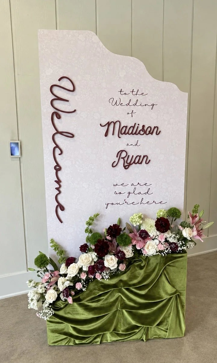 Custom burgundy acrylic wedding welcome sign with three personalized wooden letters, lush floral arrangements, and olive green draping, designed and built by Covenant Design Co. for an event at Bella Cavalli in Aubrey, Texas.