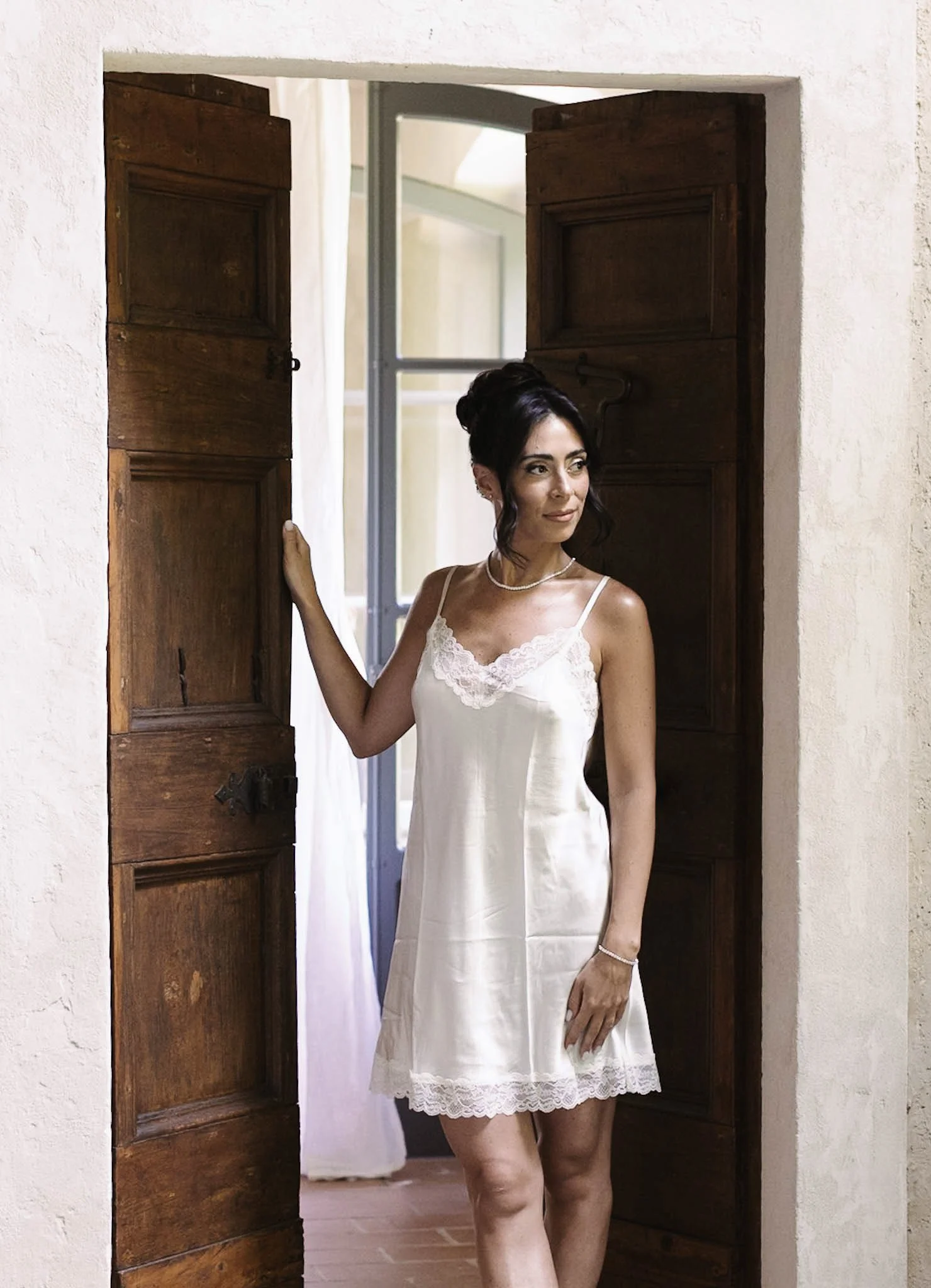 Una donna in vestaglia di seta bianca si trova in una porta di legno, in una stanza luminosa con pareti beige e una finestra aperta.