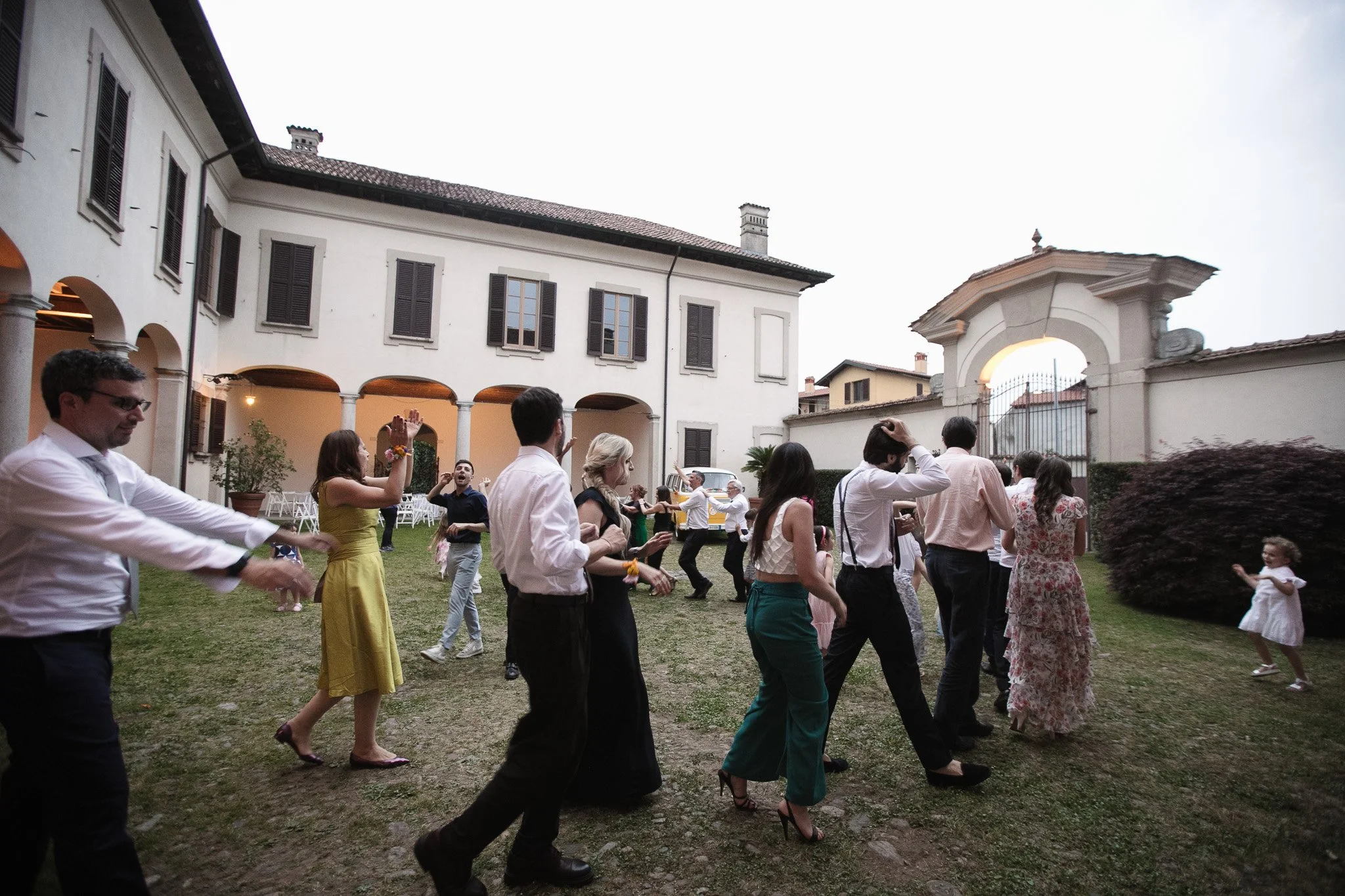 Persone che ballano e si divertono nel cortile di una casa storica, con un arco d'ingresso e un edificio con finestre in background, durante un evento sociale.