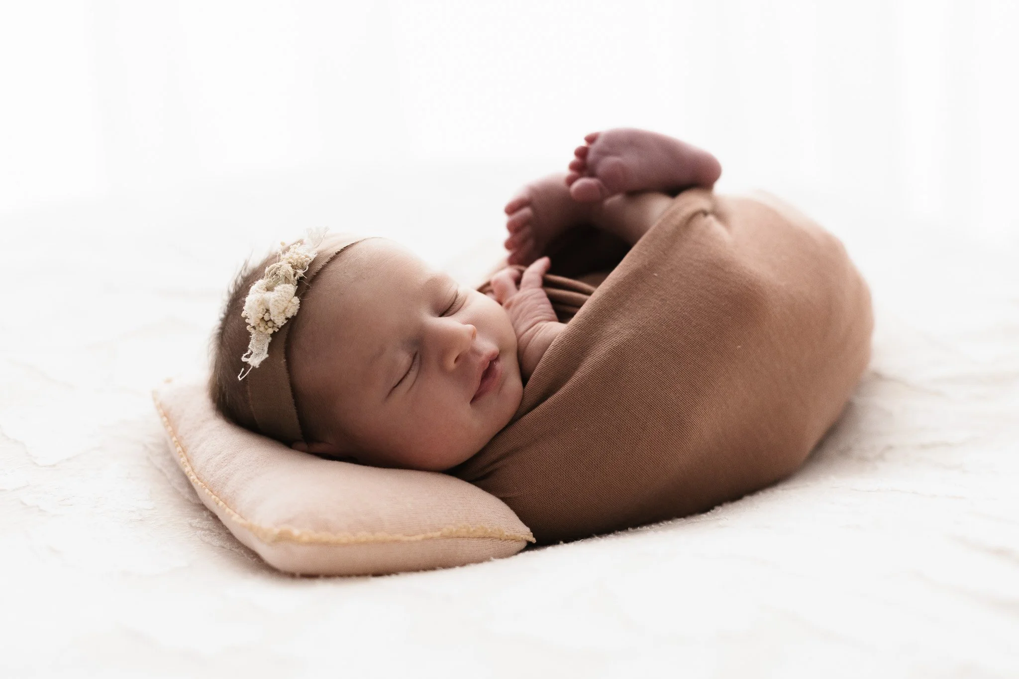 Neonato sdraiato su un cuscino, avvolto in una copertina marrone, con una fascia floreale sulla testa, mentre dorme pacificamente.