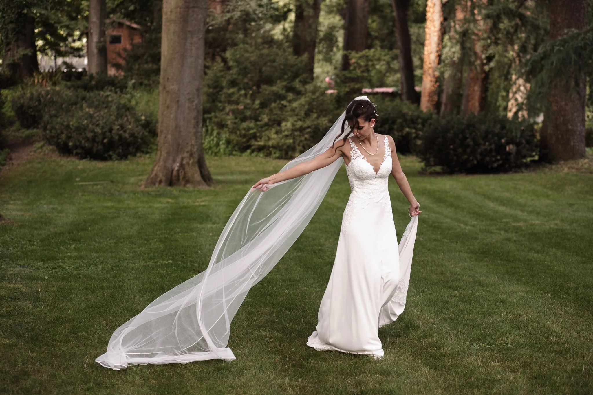 Una sposa in abito da sposa bianco con velo lungo si trova su un prato verde in un parco, circondata da alberi.