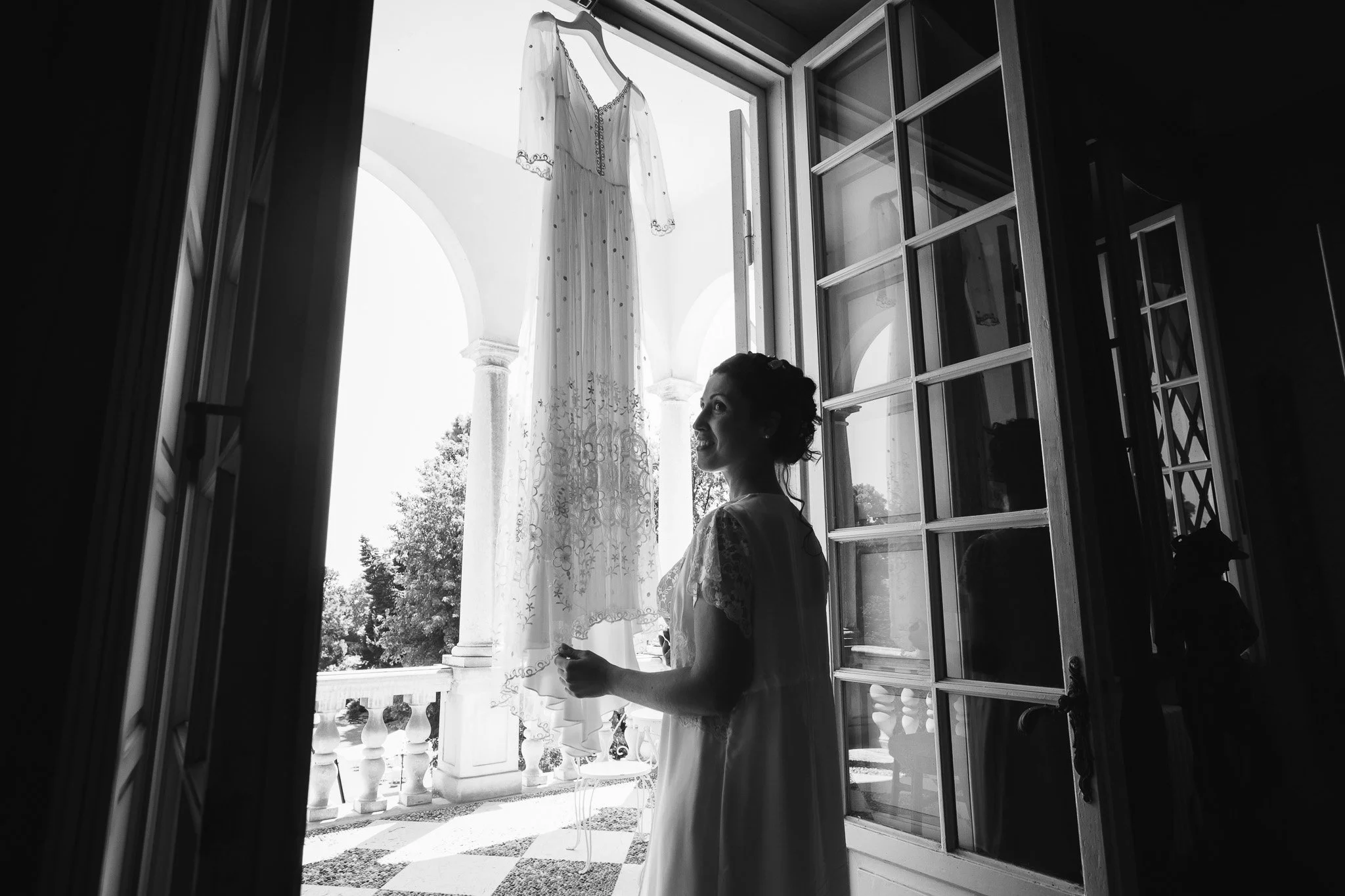 Una sposa sorride guardando una gonna nera appesa all'esterno di un balcone, in una stanza con porte e finestre aperte in stile classico