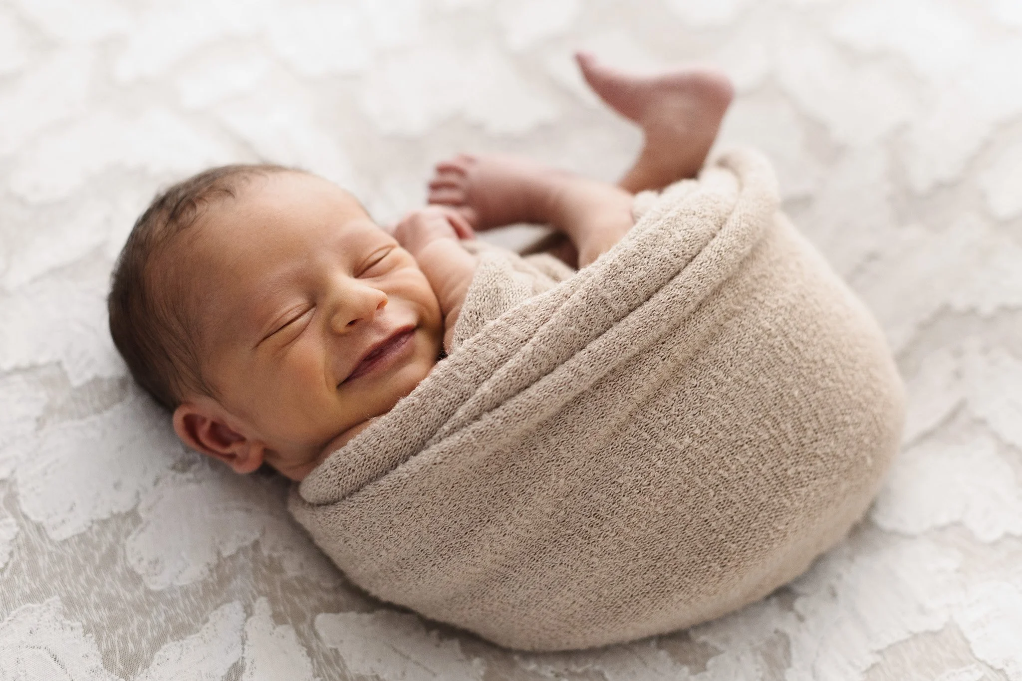Nascita di un neonato avvolto in una coperta beige, sdraiato su un letto con tessuto ricamato bianco, che sorride felice.