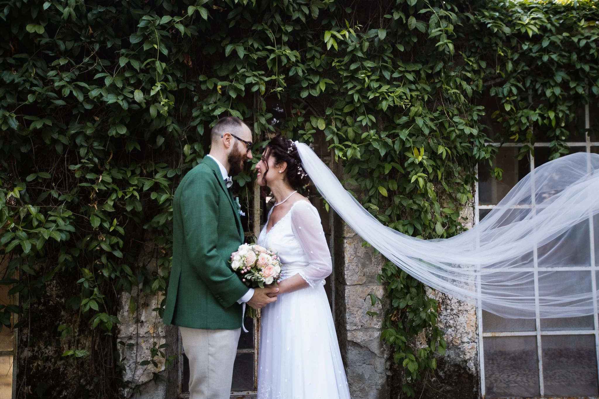 Una coppia di sposi si bacia davanti a un muro coperto di edera, la sposa indossa un abito bianco con un velo lungo e tiene un bouquet di fiori rosa e bianchi, l'uomo indossa un abito verde. Sono molto vicini, con il loro viso quasi a toccarsi.