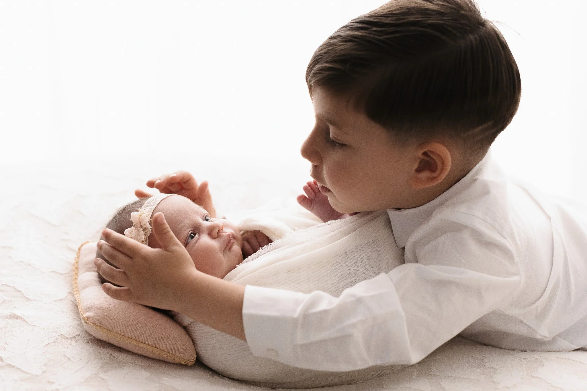Un bambino giovane tiene in braccio una neonata, guardandola con affetto, in un'illuminazione morbida e uno sfondo bianco.