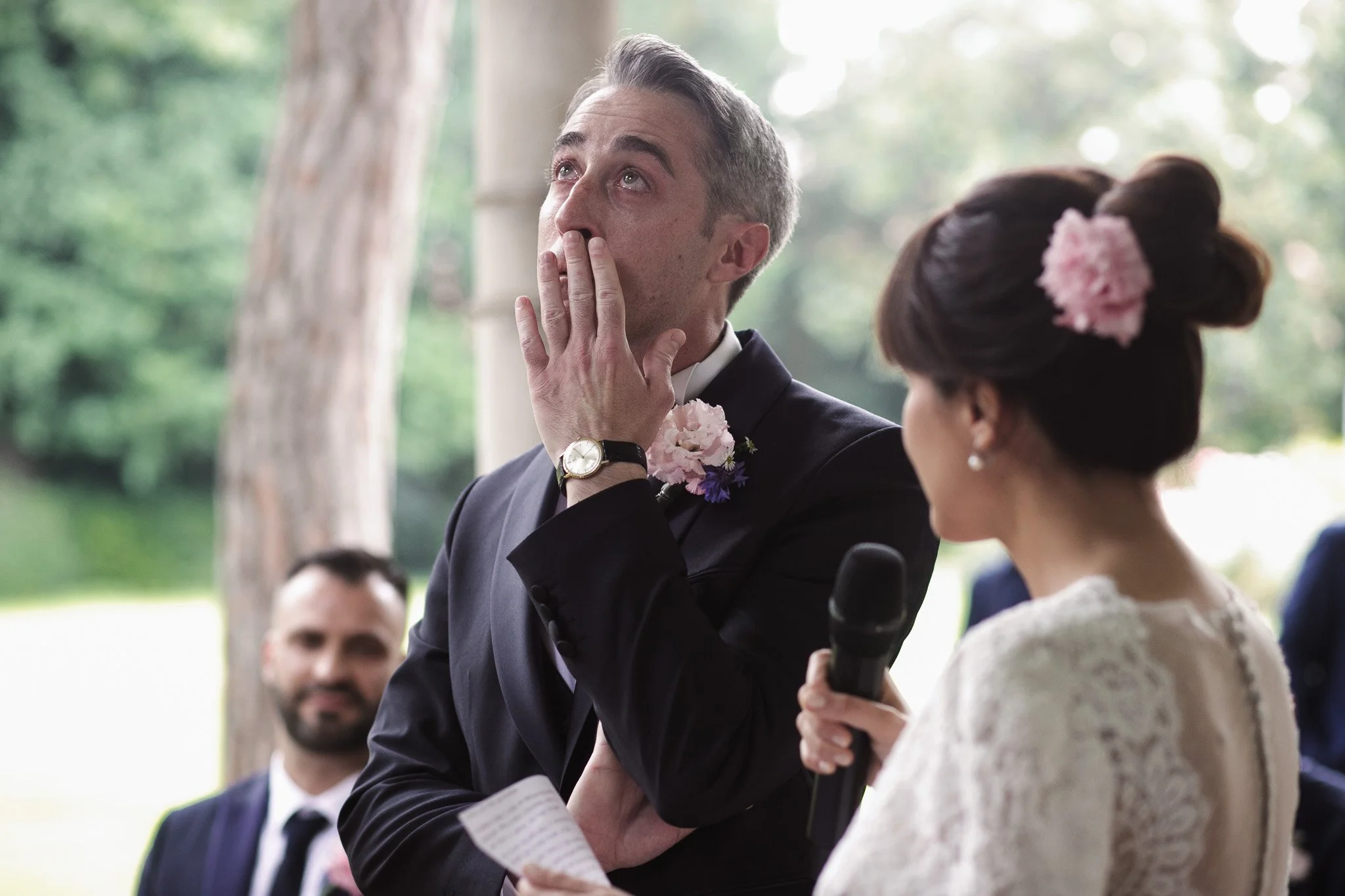 Un uomo sta piangendo commosso durante un matrimonio, mentre una donna con un fiore rosa tra i capelli gli tiene il braccio e ha in mano un microfono. Un altro uomo sorride sullo sfondo.