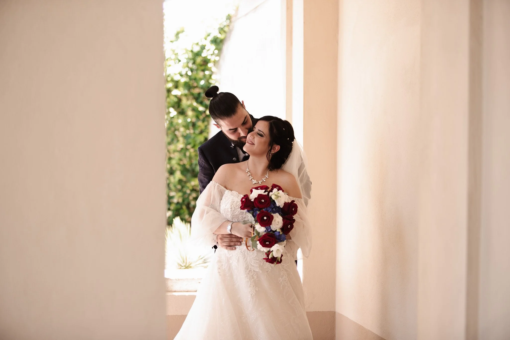 Una coppia di sposi si abbraccia affettuosamente in un corridoio, la sposa tiene un bouquet di fiori rossi, bianchi e blu, indossa un abito da matrimonio bianco e una collana di perle, mentre l'uomo indossa un abito scuro con uno stile formale.