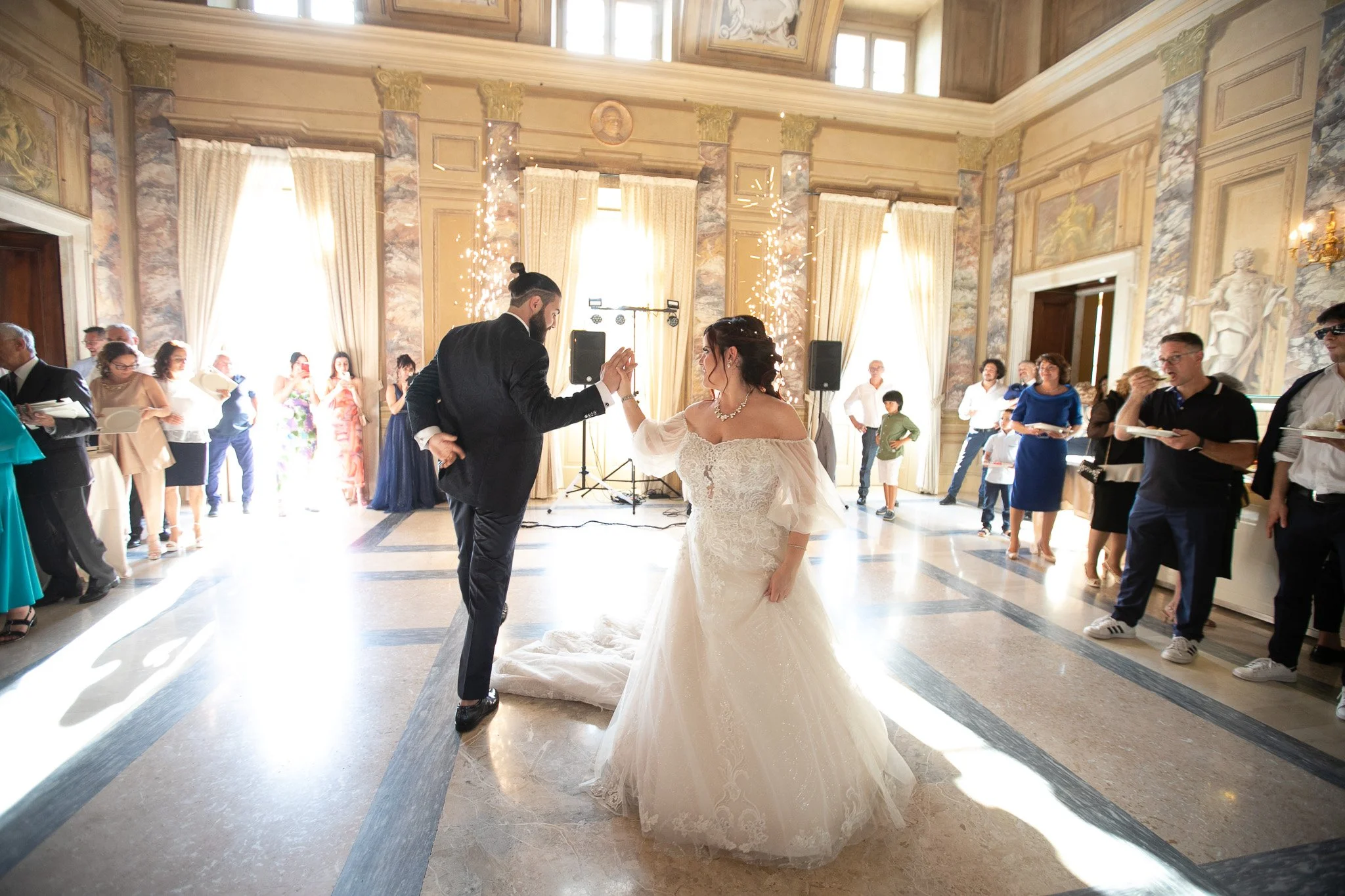 una coppia di sposi balla in una sala elegante con decorazioni classiche, accompagnata da amici e familiari; ci sono fuochi d'artificio in background