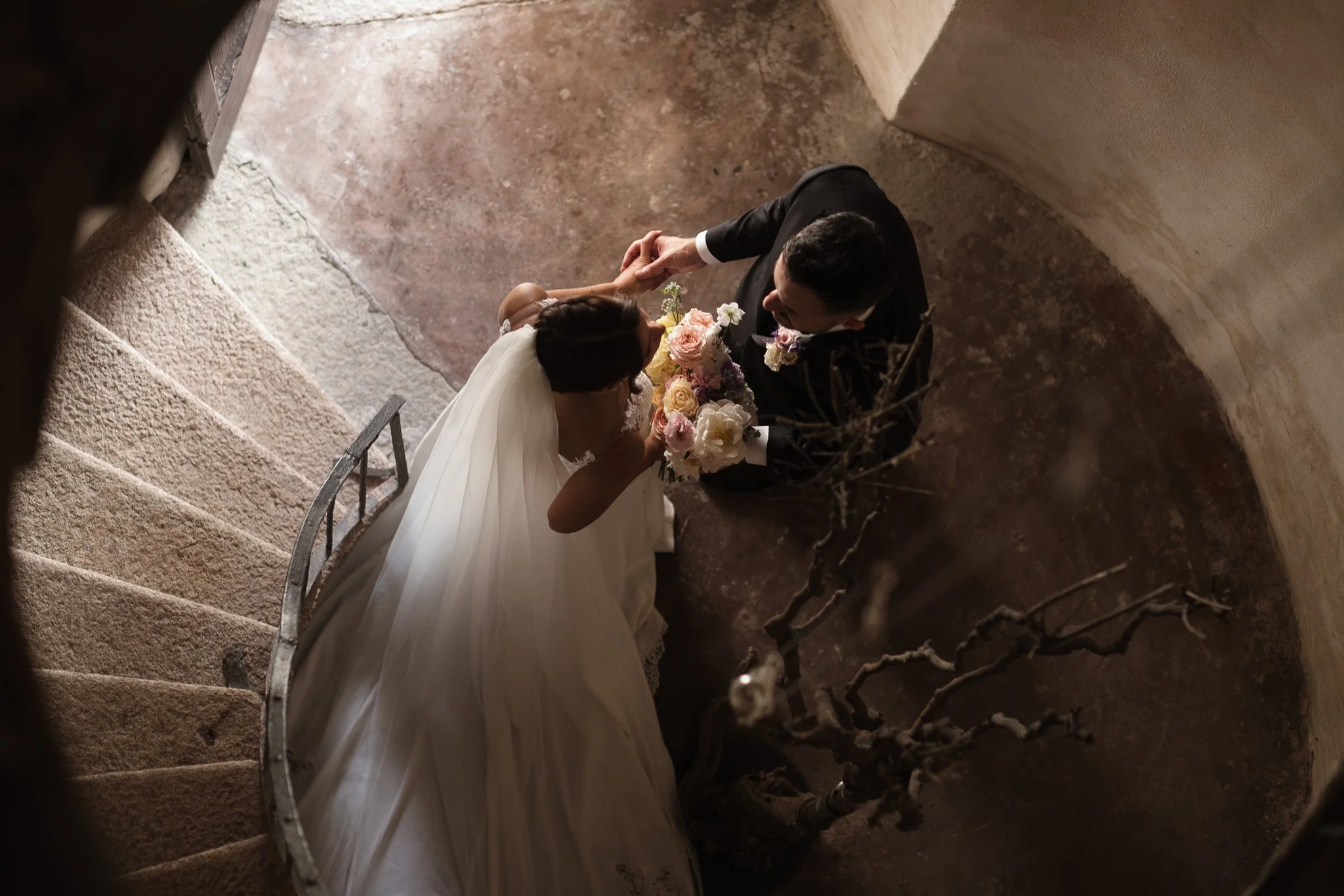 Una coppia di sposi in abito da matrimonio si abbraccia in un vicolo con pareti di pietra e un arco, con un bouquet di fiori tra di loro.